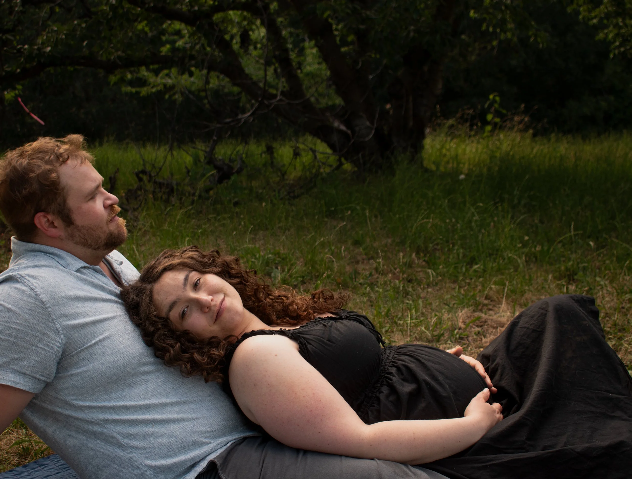 Three people lying on the grass outdoors, embracing each other, with trees in the background.