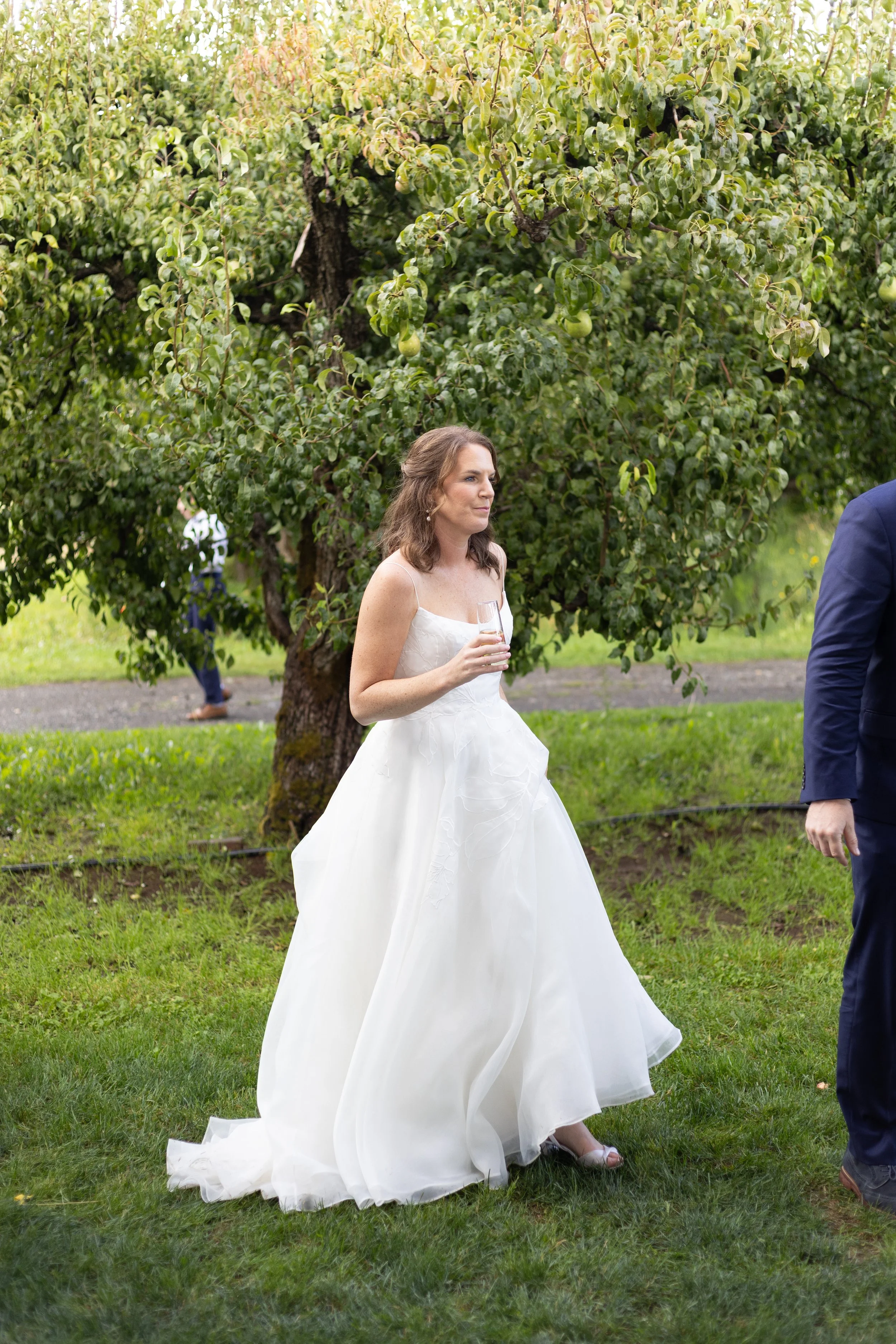 A woman in a white wedding dress holding a wine glass, standing outdoors on grass with a large tree in the background, and a man in a blue suit partially visible on the right.