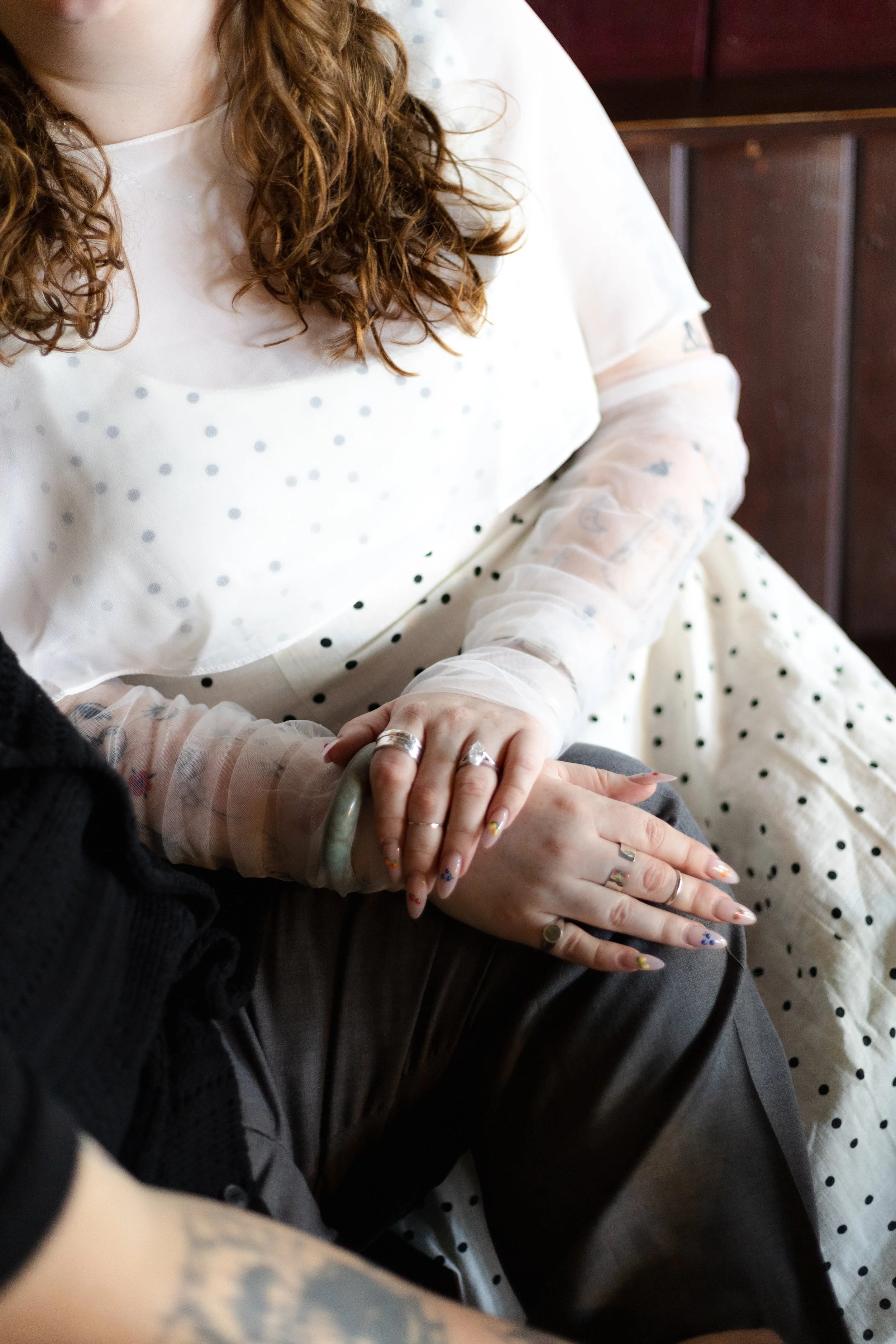 Close-up of a person with curly hair wearing a white dress with black polka dots, holding hands with another person. The person has decorated nails with various colors and patterns and multiple rings.
