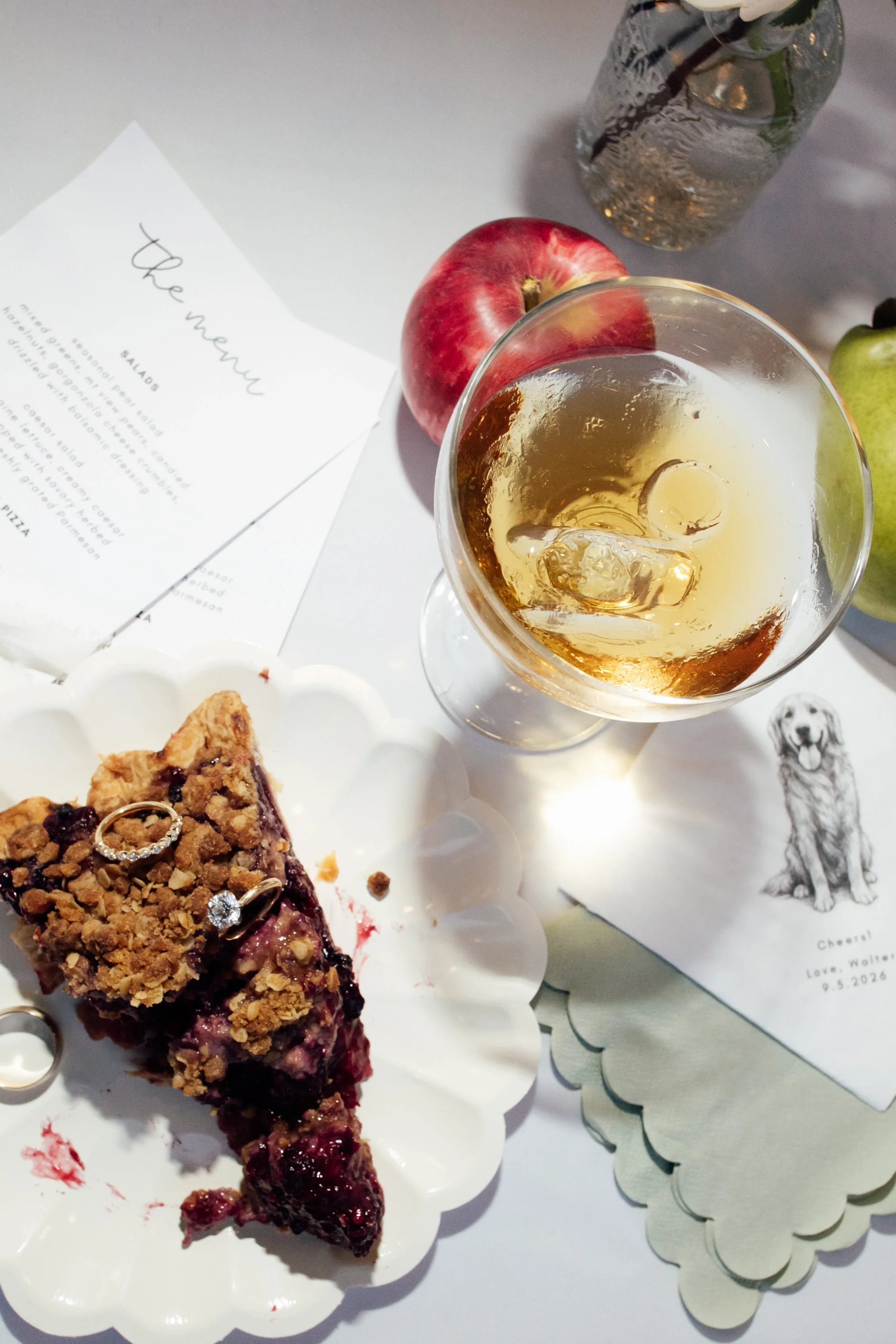 A slice of cherry crumble pie on a white decorative plate, a glass of iced drink with ice and lemon, a red apple, and a green pear. There is also a paper with a handwritten menu or note on the table, and a water bottle in the background.