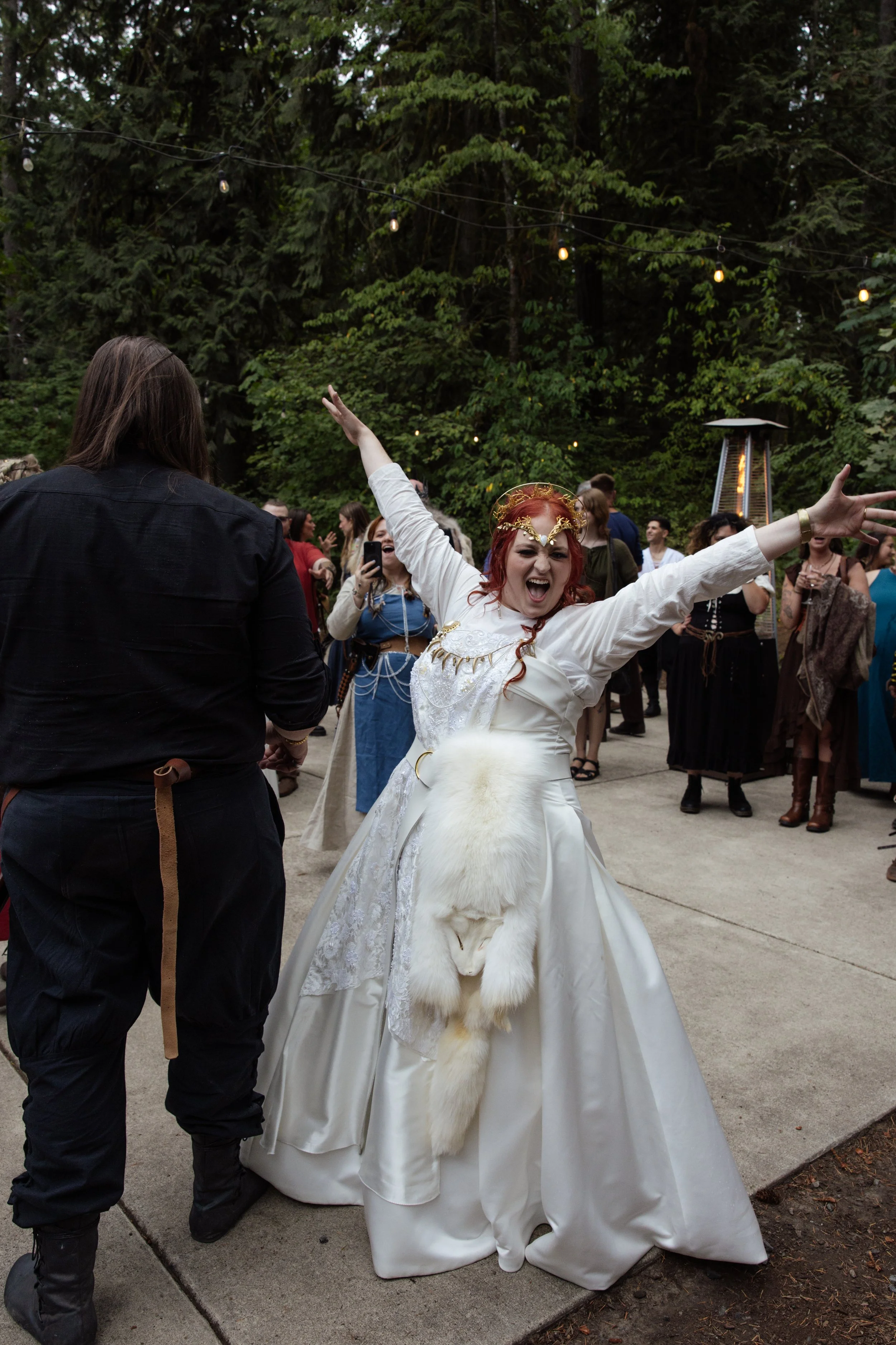 A woman dressed in a white wedding gown with a fur accent at the waist, celebrating outdoors among other people, with trees and string lights overhead.
