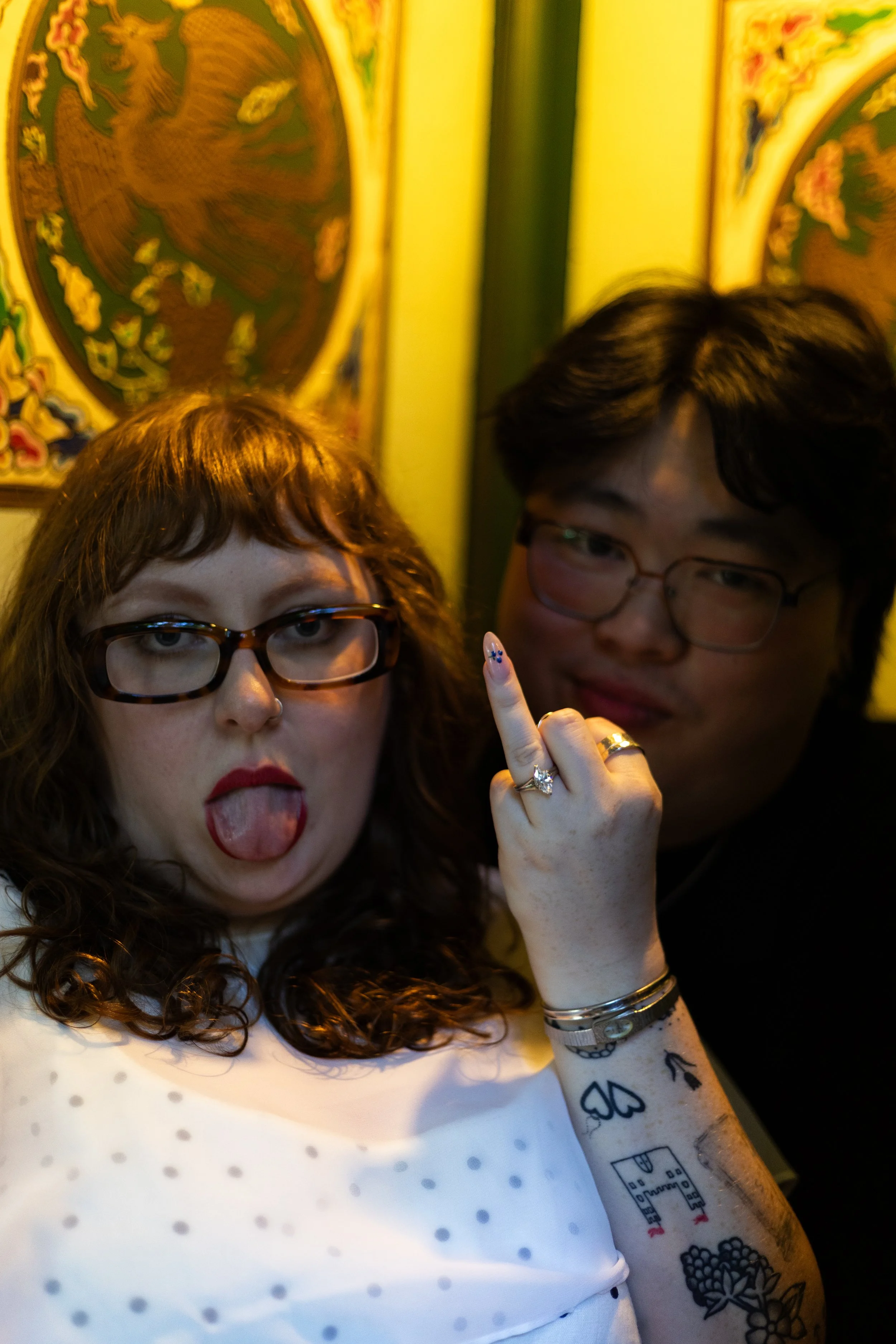 A woman with glasses sticking out her tongue, and a man with glasses making a rude gesture with his middle finger, both in front of colorful, traditional Asian artwork.