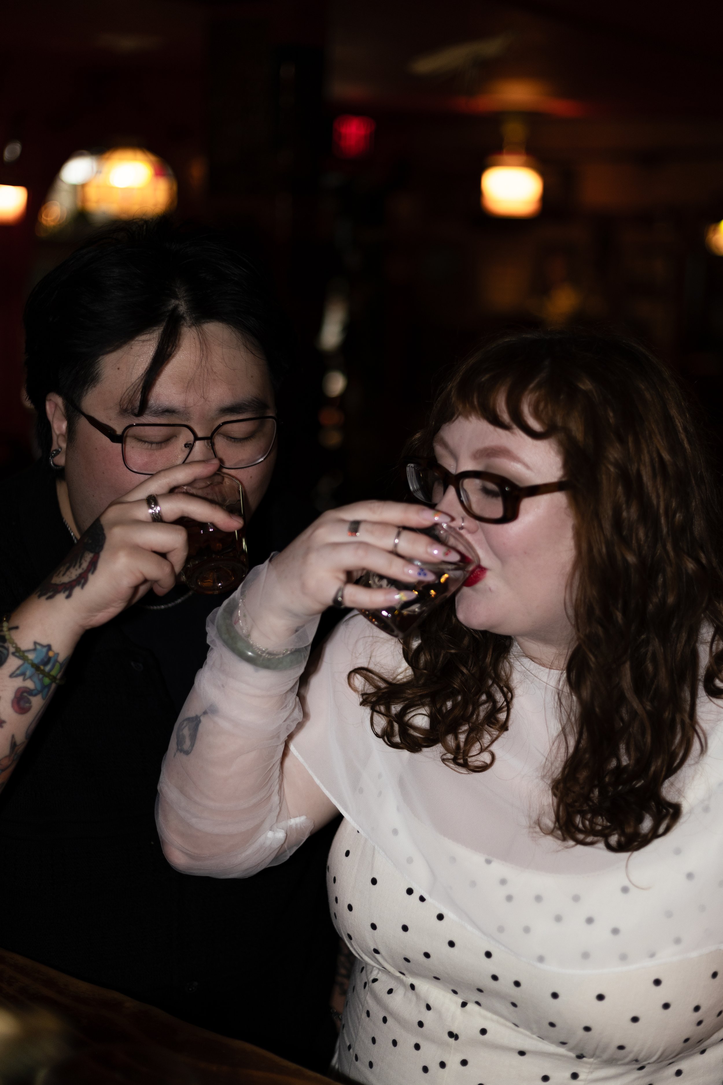 Two women with glasses and tattoos drinking from glasses during a social gathering indoors at night.