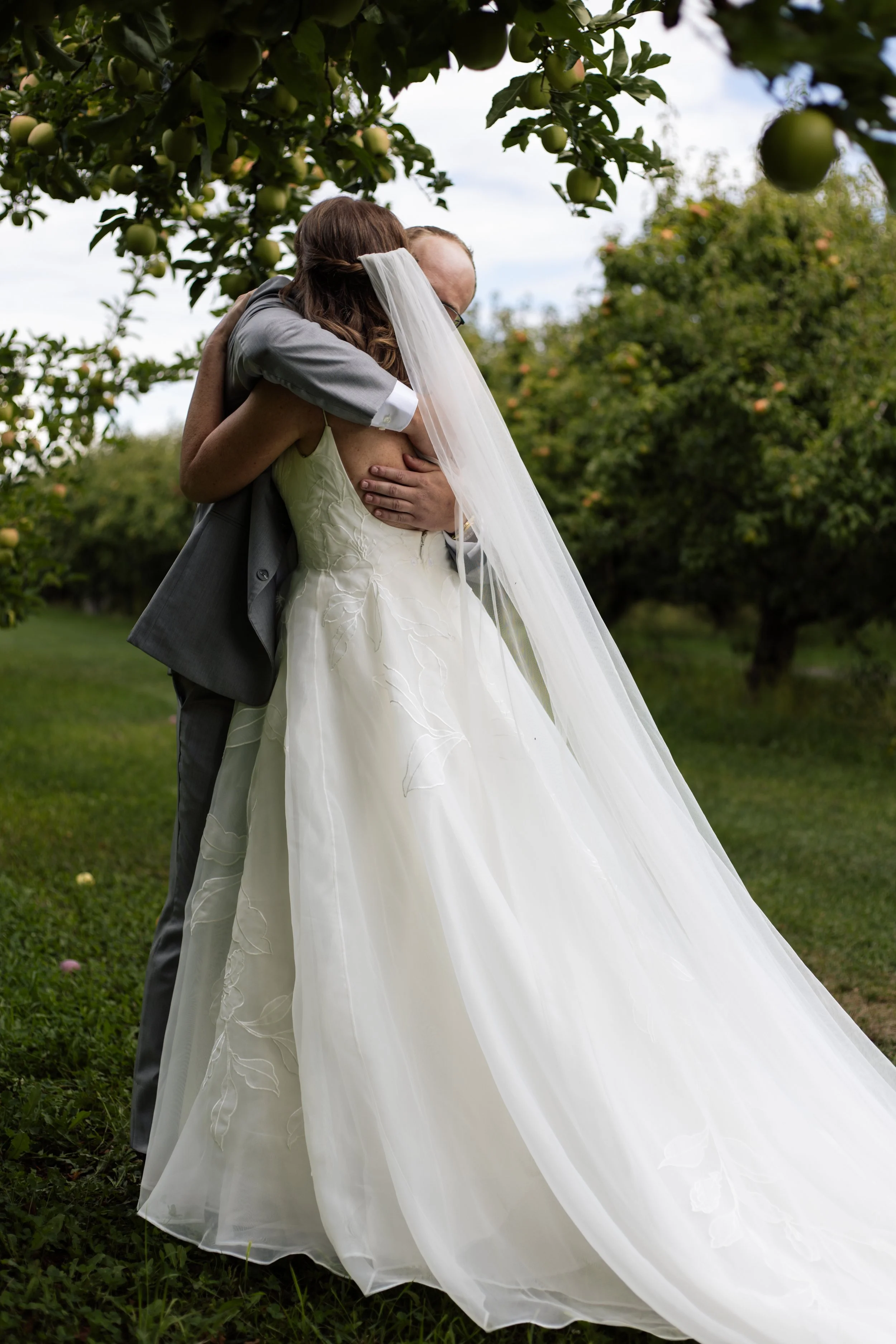 Couple hugging outdoors during a wedding, woman in a white wedding dress and veil, man in a gray suit, green trees and grass visible.