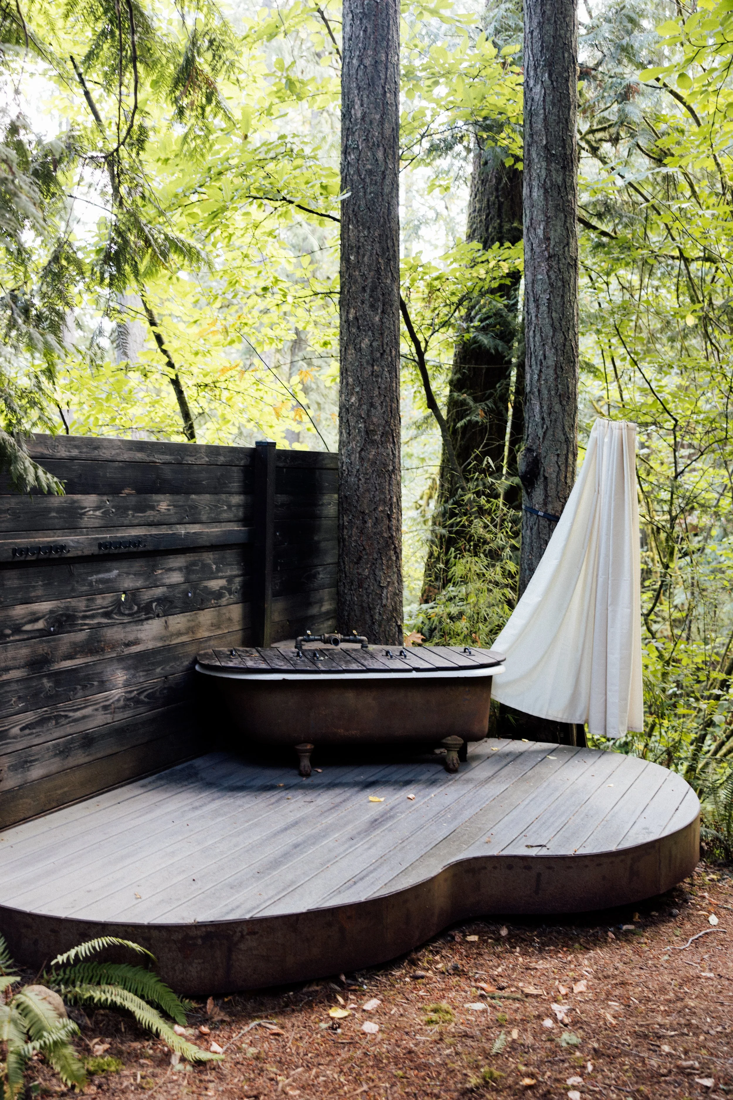 A rustic outdoor shower setup with a clawfoot bathtub, a wooden deck, a black wooden privacy fence, and a white shower curtain amidst a forest of tall trees and lush green foliage.