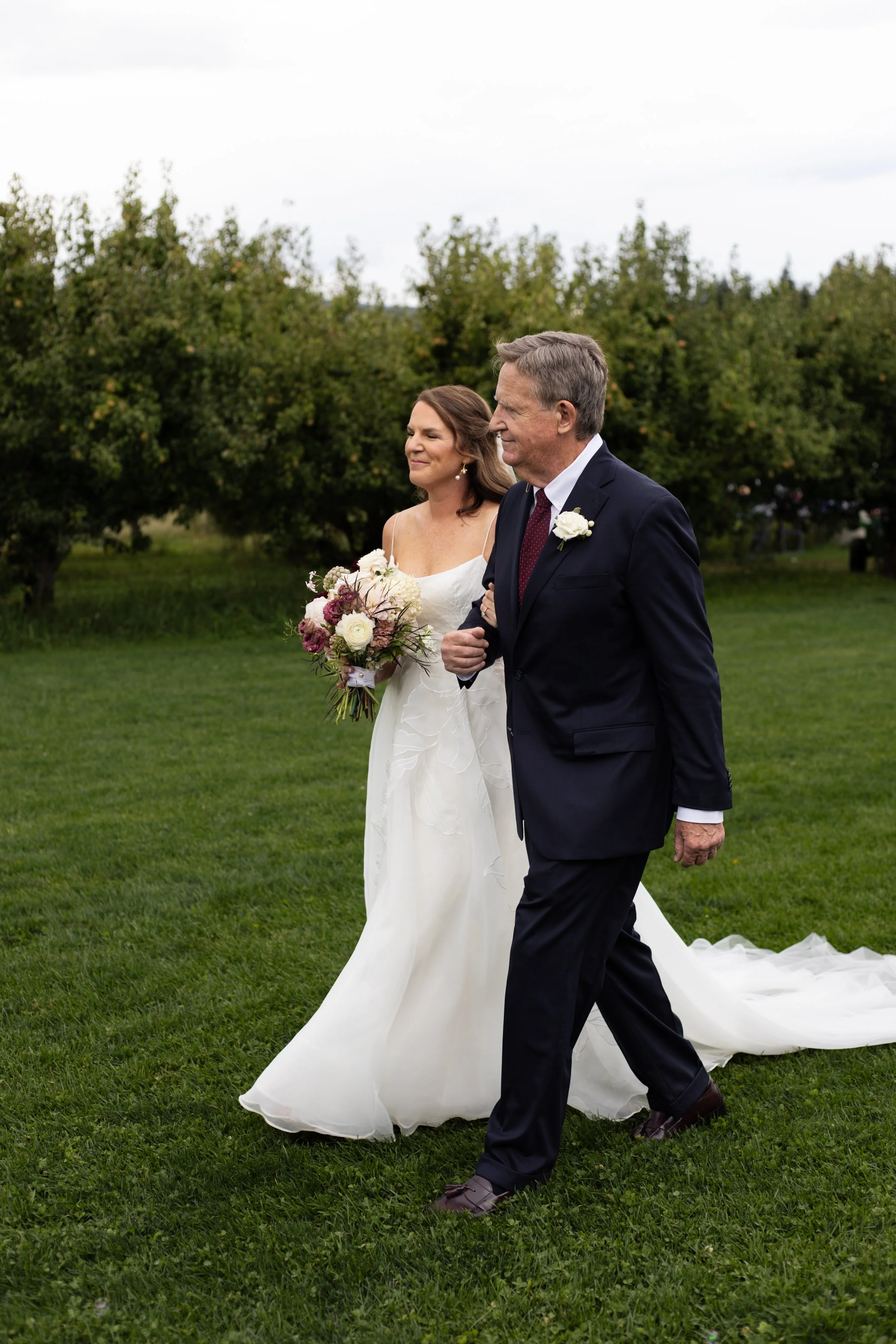 A bride in a white wedding dress holding a bouquet walking with an older man in a dark suit outdoors during the daytime, with trees and a cloudy sky in the background.