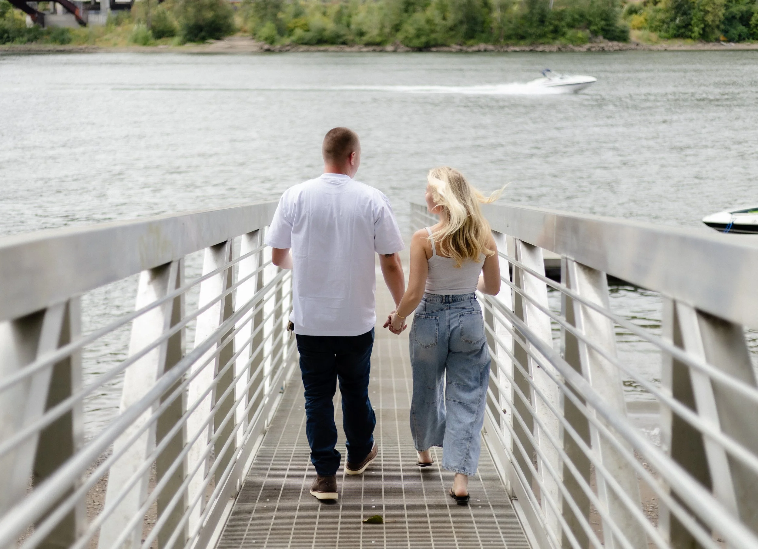 A couple holding hands walking on a dock by the water with a boat speeding past in the background.