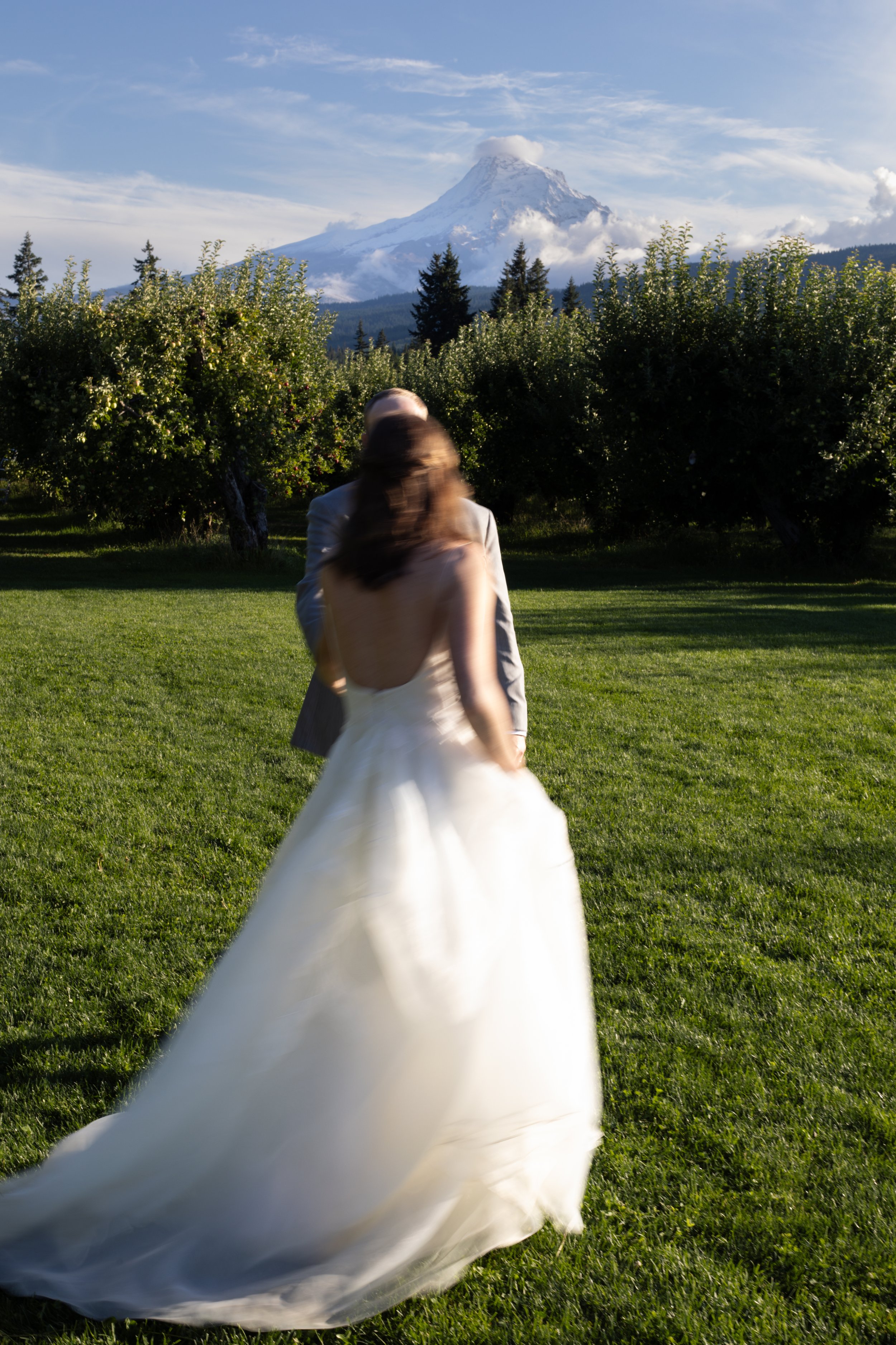 A bride in a white wedding dress dancing with a groom in a gray suit outside on green grass, with trees and a mountain with snow in the background.