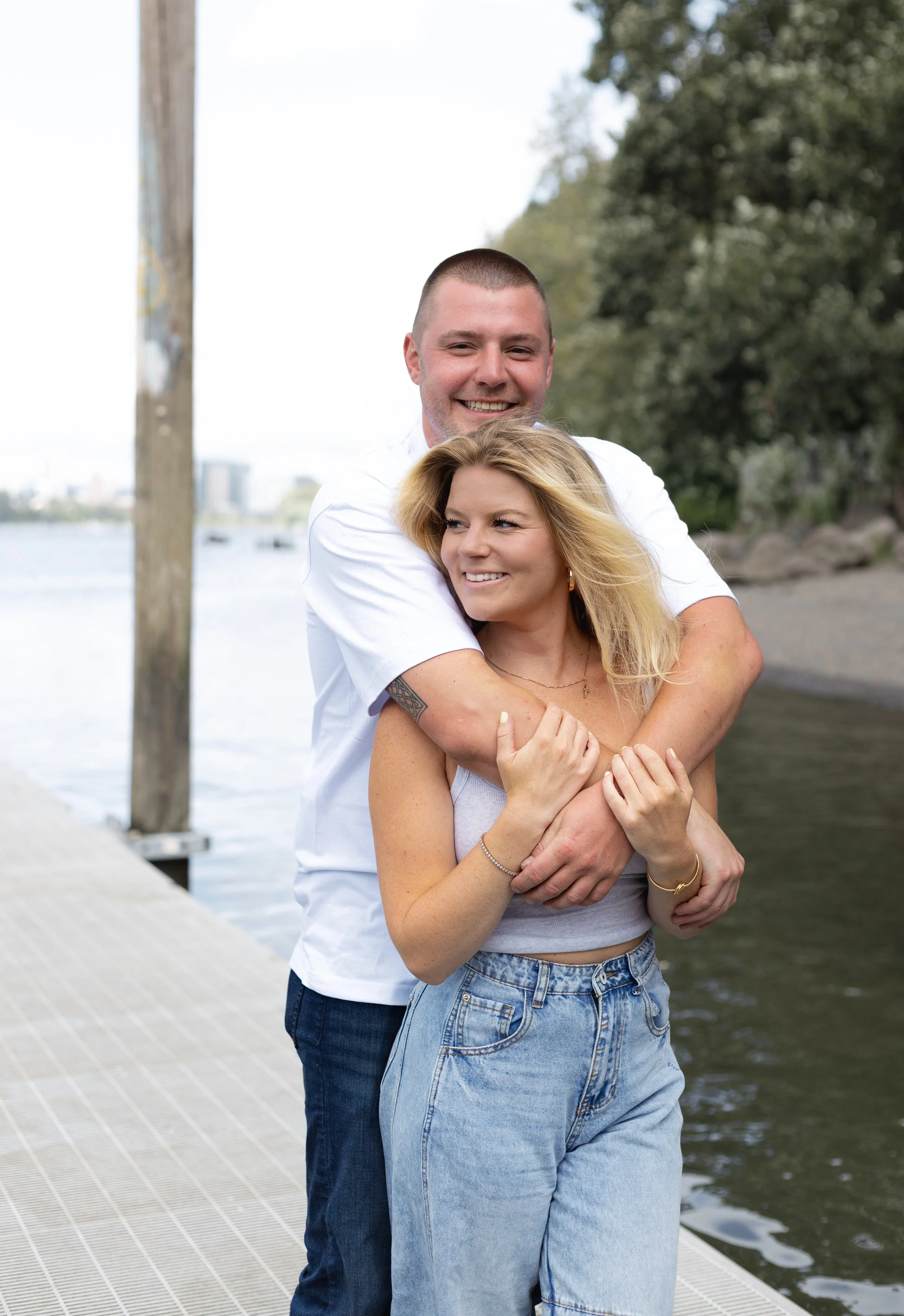 A happy couple hugging by the river on a cloudy day, with trees and city buildings in the background.