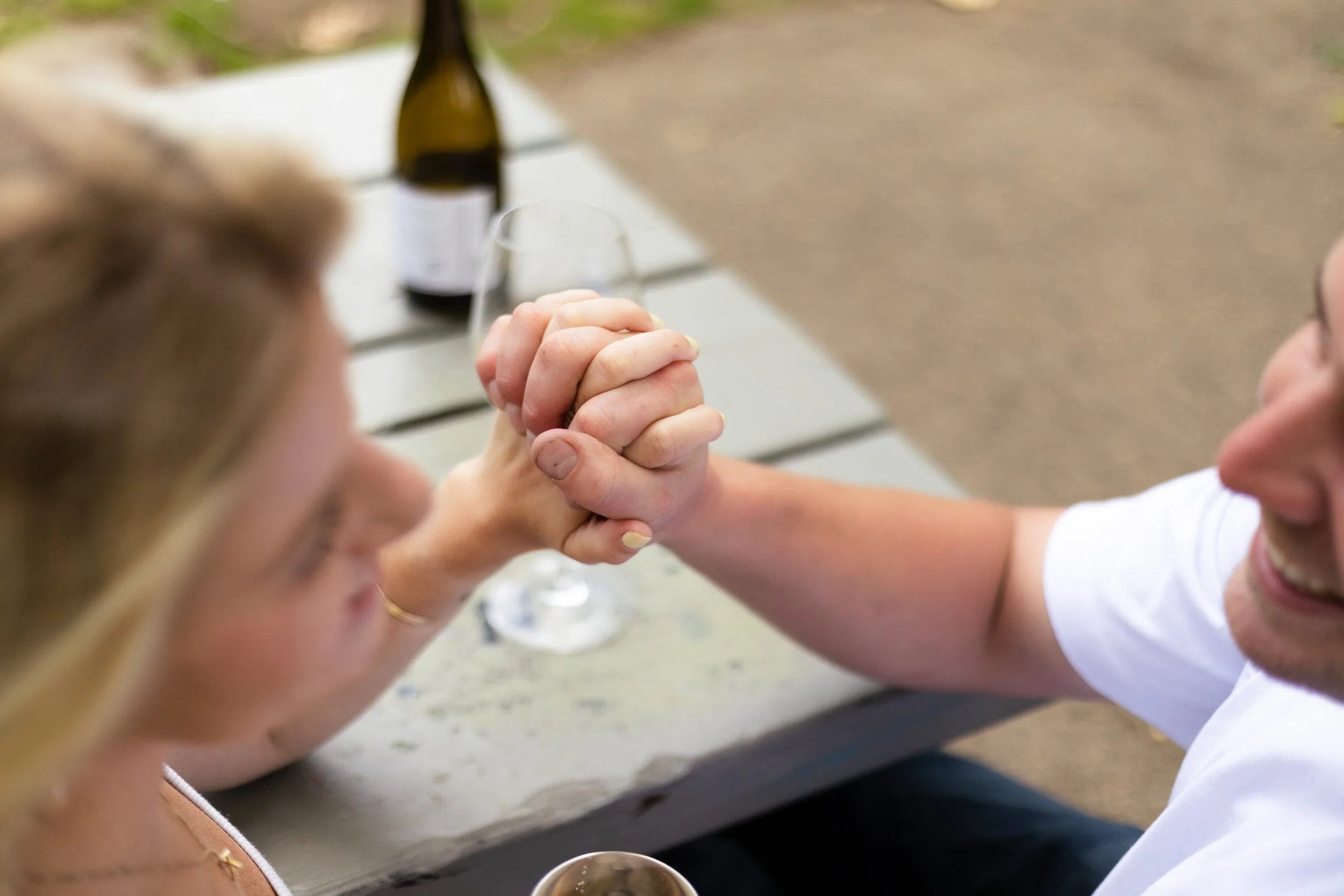 Two people arm wrestling at a table outdoors, with one person's face partially visible, and a bottle and a glass in the background.
