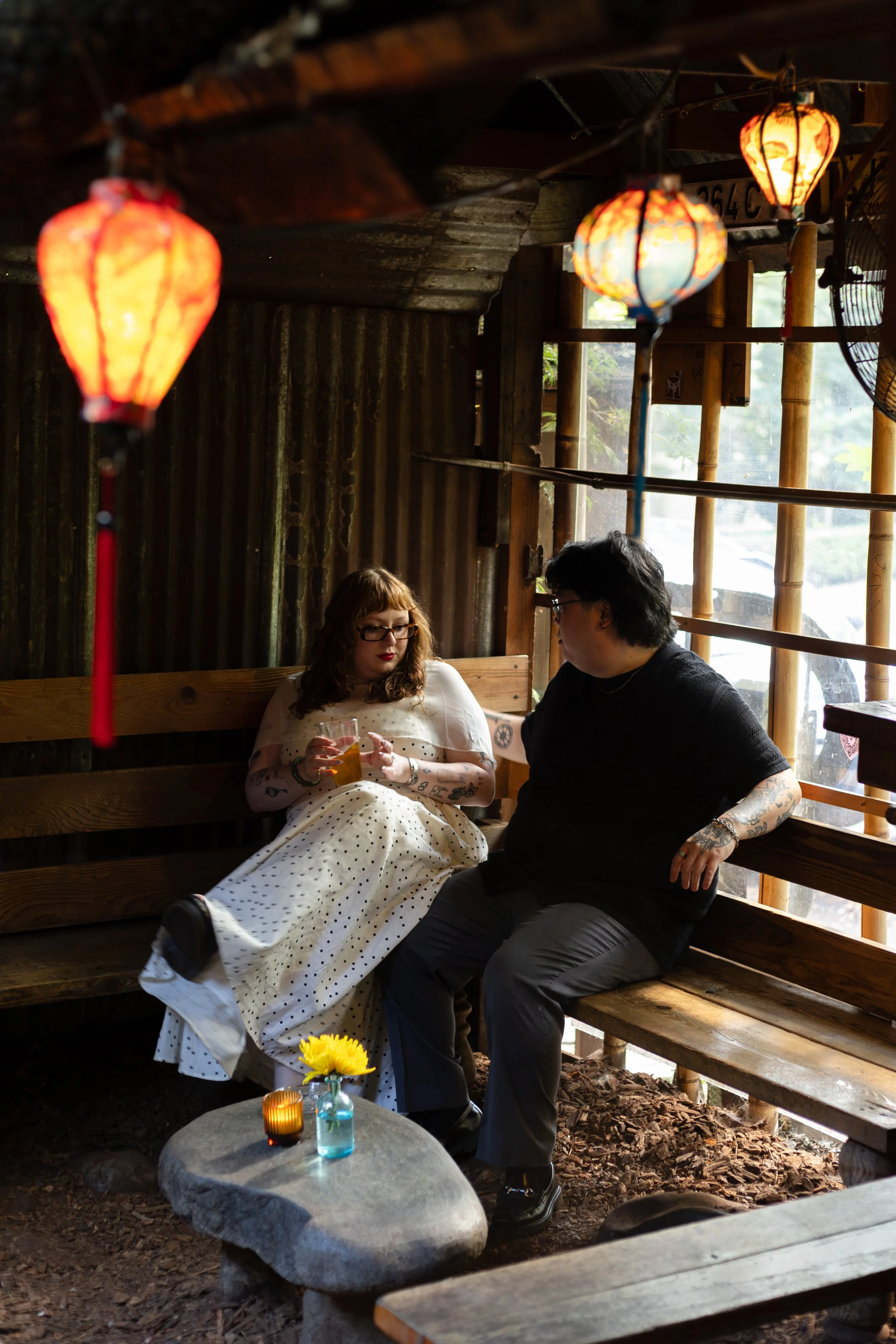 A woman in a white polka dot dress and a man in a black shirt sitting on a wooden bench inside a rustic structure with hanging lanterns, a window, and decorative items on the table.