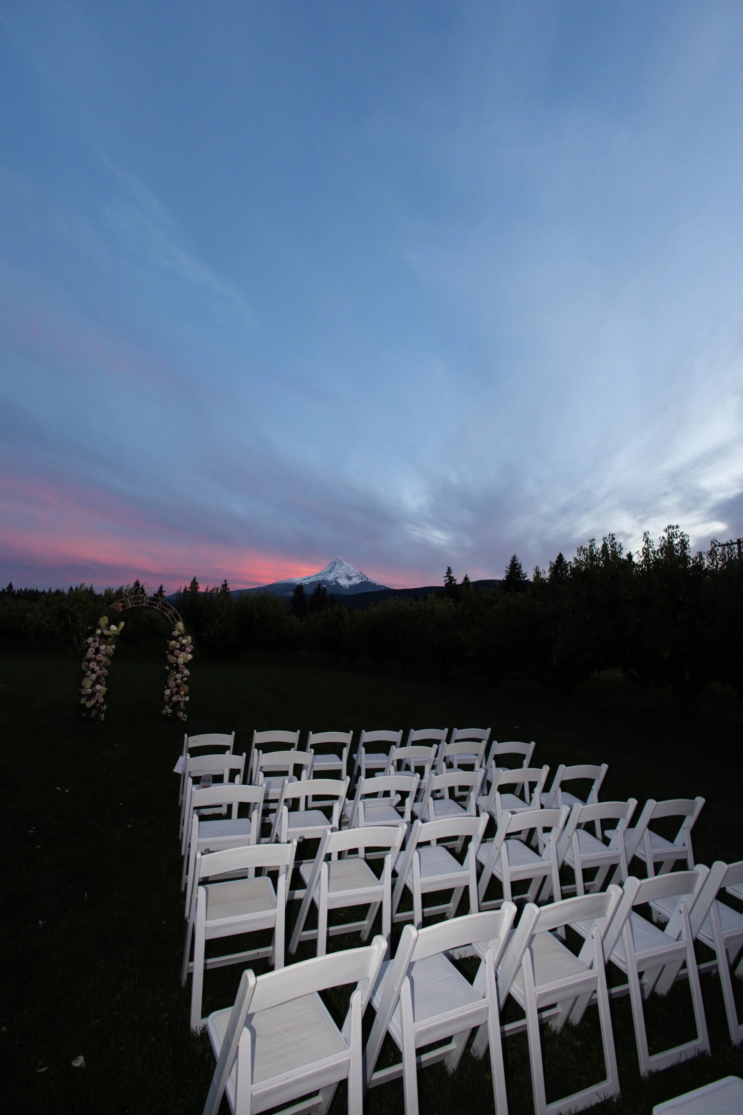 White chairs arranged in rows outdoors facing an arch decorated with flowers, with a mountain and sunset sky in the background.