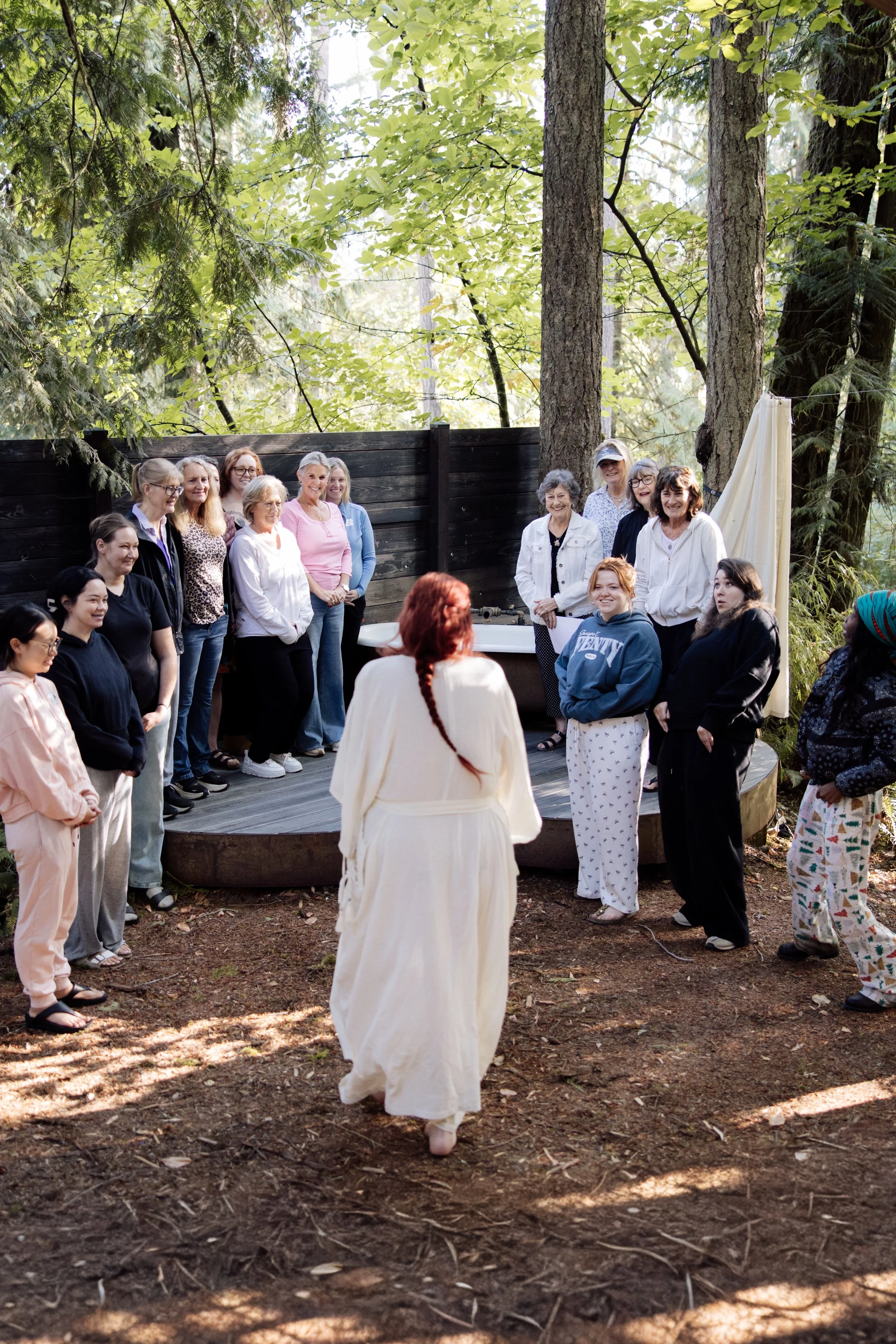 A group of women standing and sitting outdoors in a wooded area with tall trees and green foliage, gathered for a social event or celebration. One woman in the foreground has red hair and is wearing a long white dress, facing away from the camera.