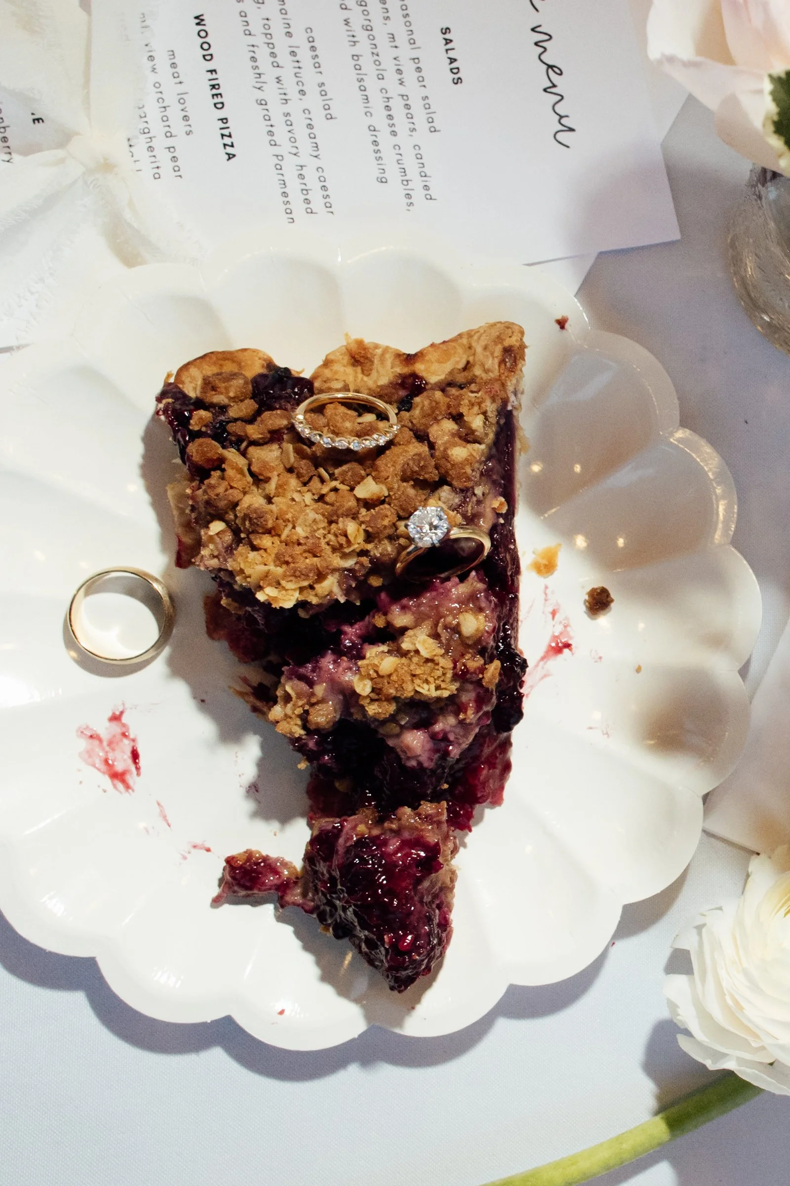 A slice of berry dessert with a crumb topping, sliced on a decorative white plate. Two rings rest on top of the dessert, and a menu is partially visible in the background.