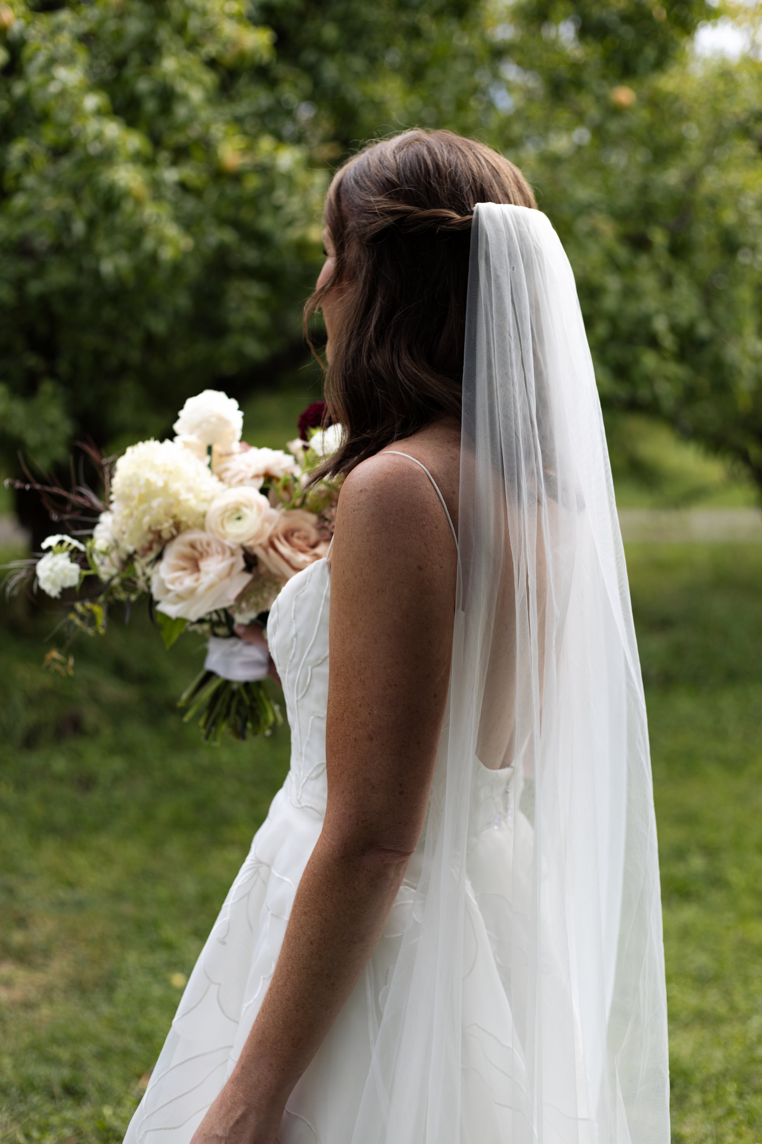 Back view of a bride in a white wedding dress holding a bouquet of flowers outdoors with a background of green trees.