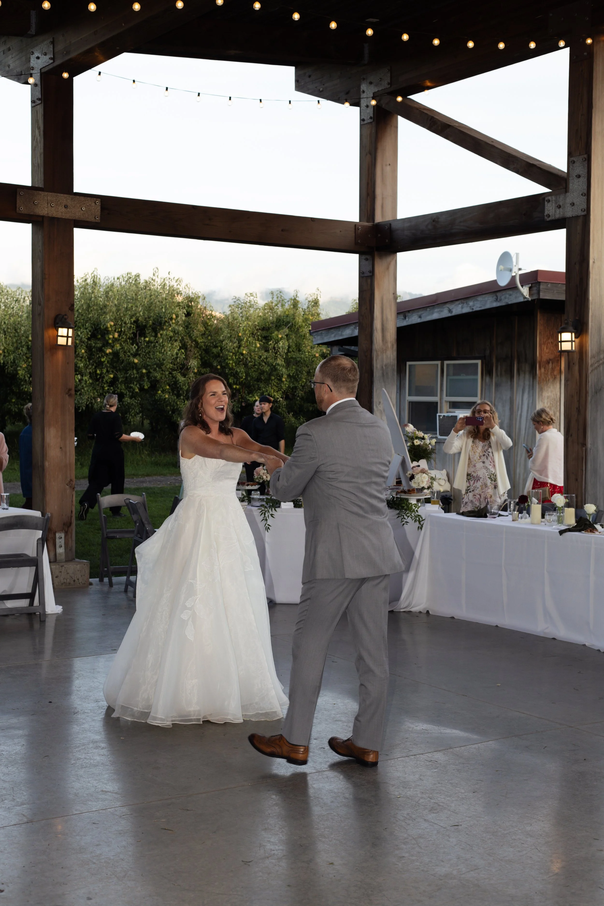 A bride and groom are dancing at their wedding reception under a covered outdoor wooden structure decorated with string lights. The bride is wearing a white wedding gown and the groom is in a light gray suit. Several guests are in the background, som