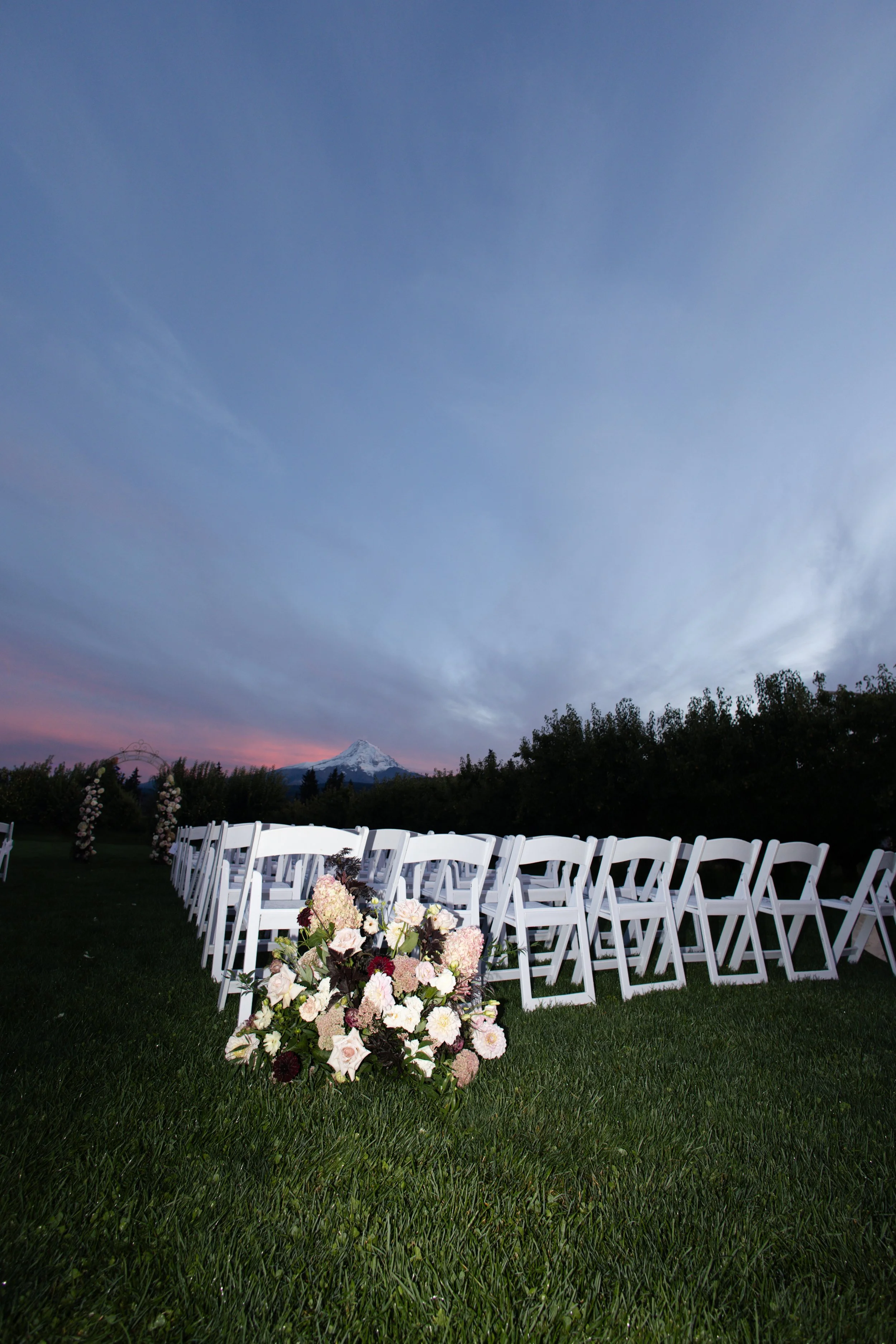 Outdoor wedding setup with white chairs arranged in rows on grass, floral arrangements in front, and snow-capped mountain in the background under a partly cloudy sky.
