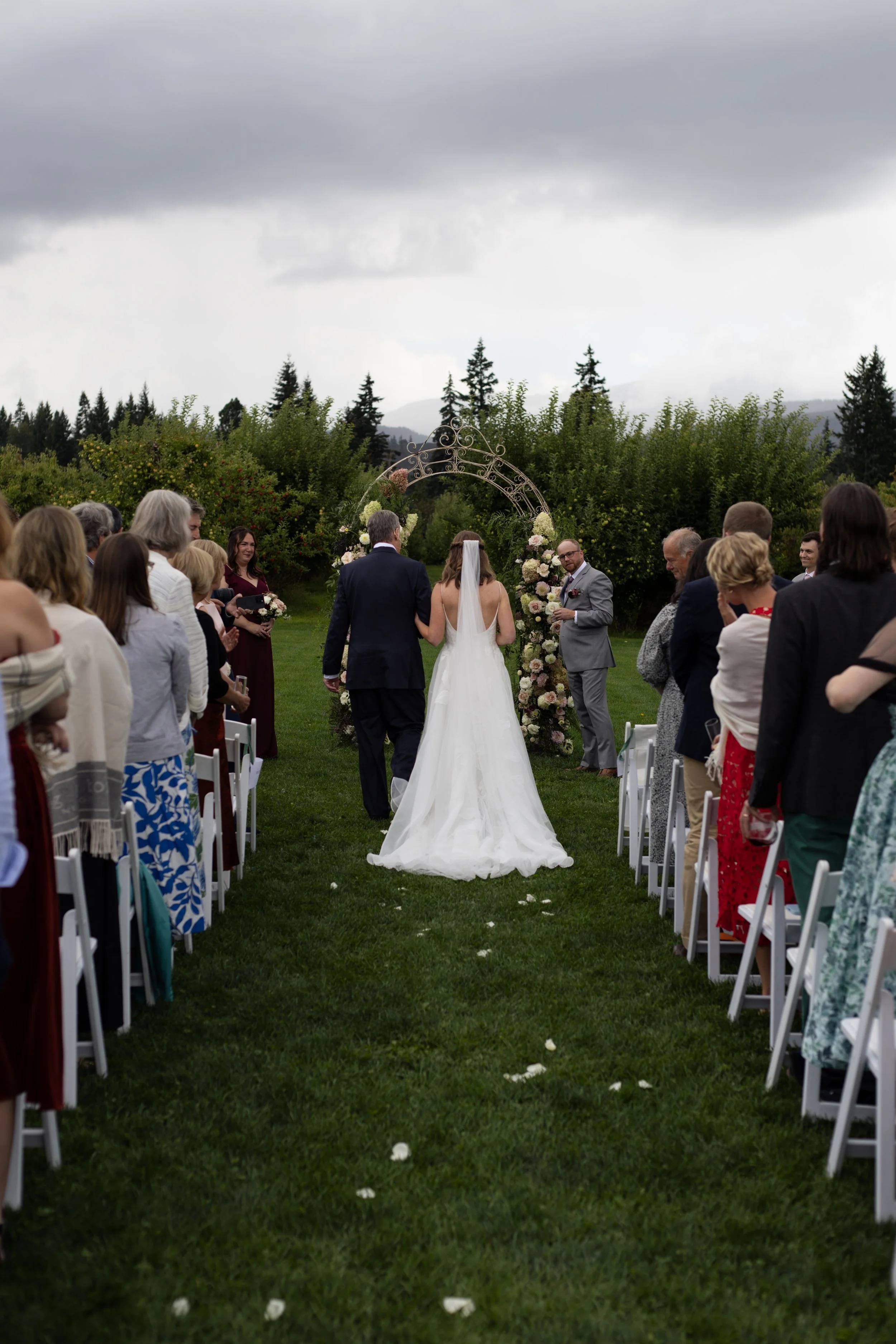 Outdoor wedding ceremony with a bride and groom facing an officiant, guests seated on either side, and a floral arch in a garden setting with Mt. Hood in the background at Mt. View Orchards