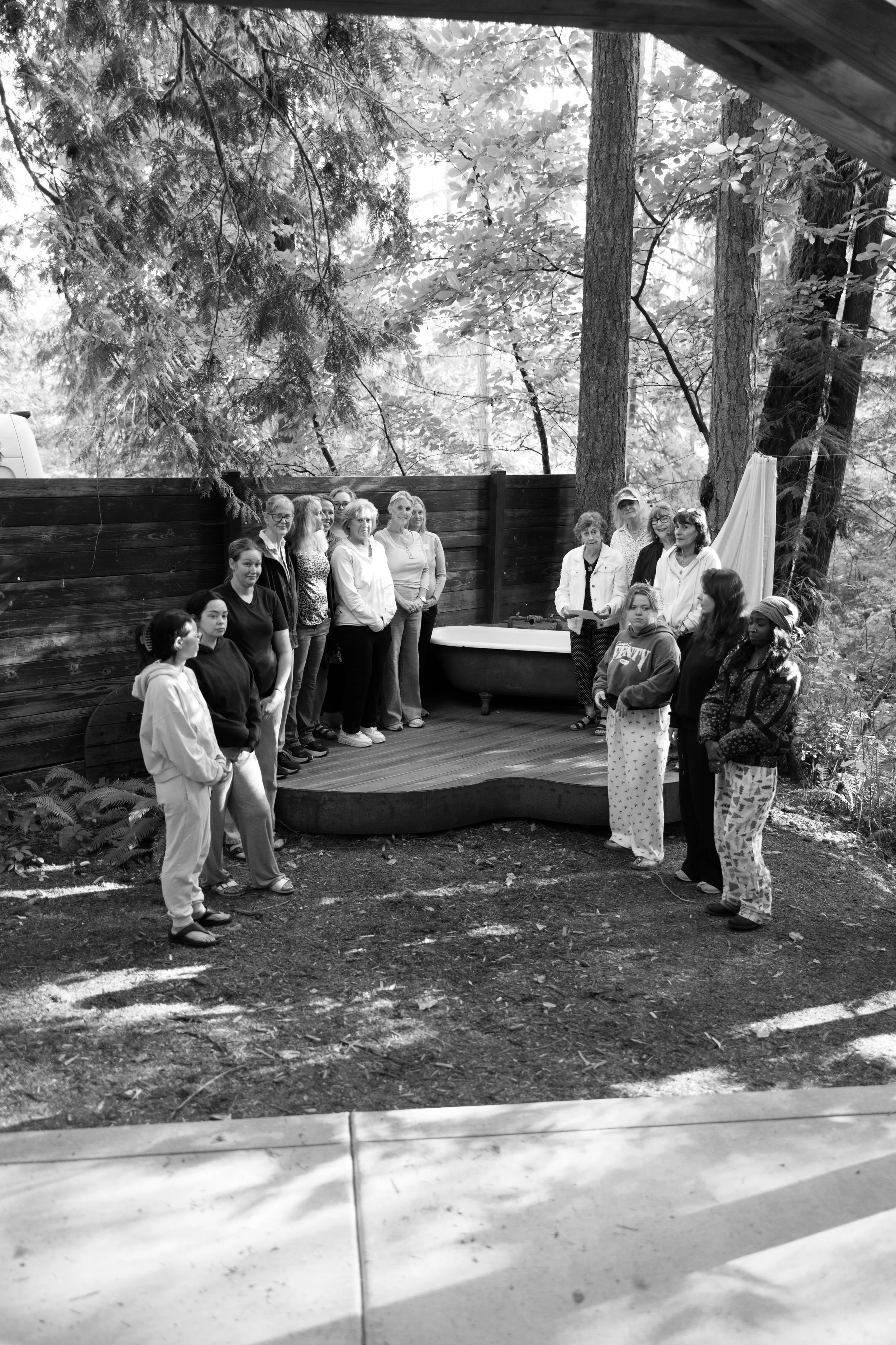 A group of people, mostly older women, standing outdoors in a semi-circle around a woman who appears to be leading or speaking. The setting is a natural environment with tall trees and a wooden fence in the background, with shade patterns on the grou
