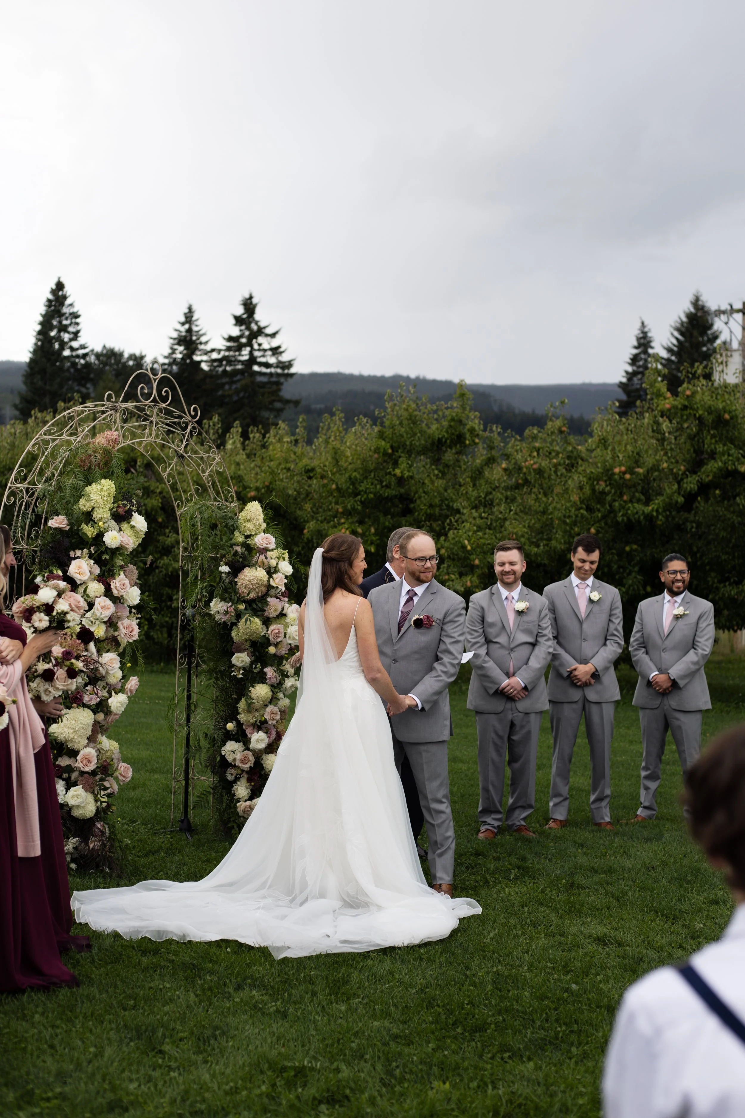 A bride and groom stand outside under an archway decorated with flowers, during their wedding ceremony, with attendants nearby and a natural landscape in Mt. View Orchards in Hood River, Oregon