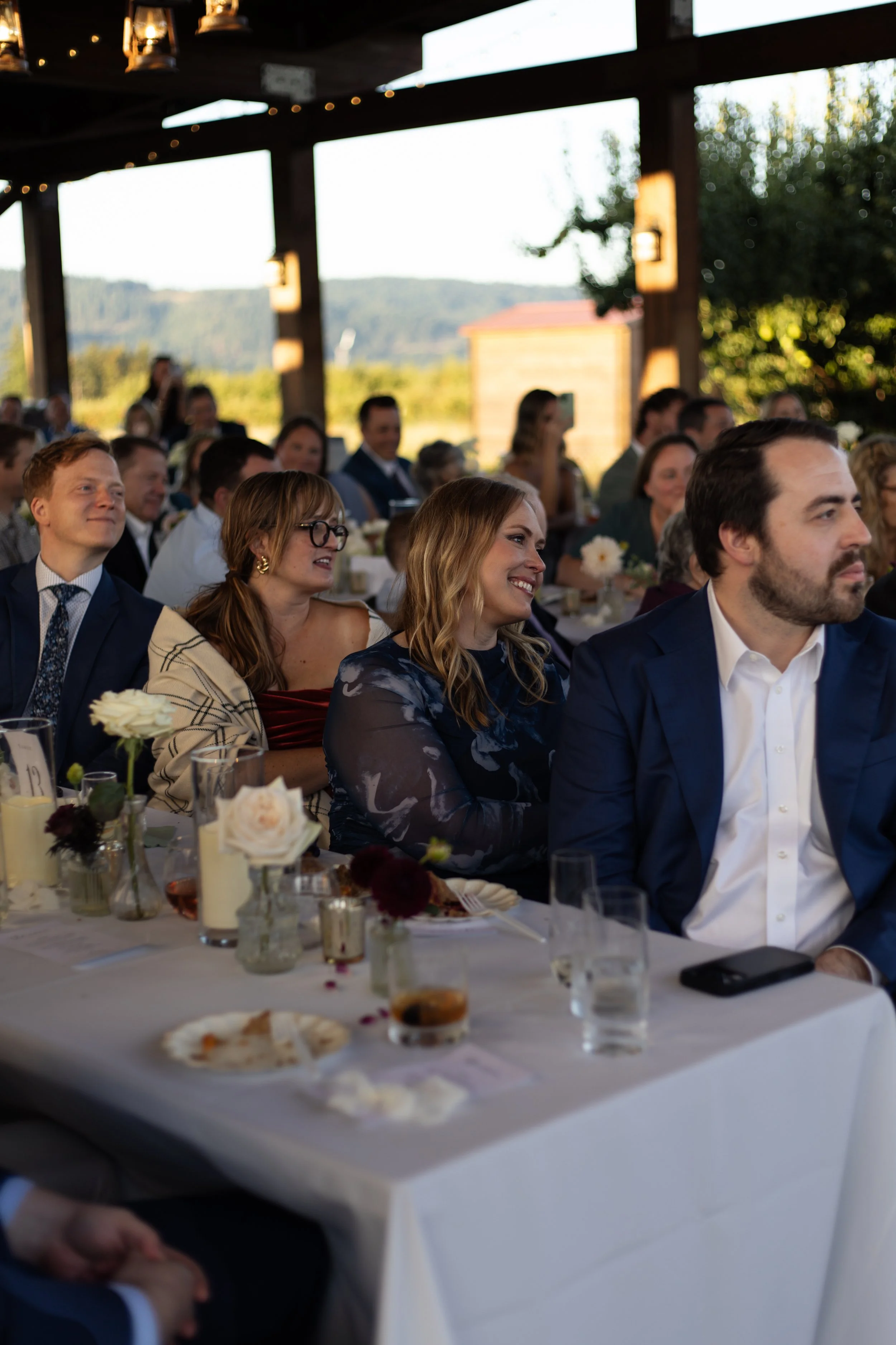 People attending a celebration or event, sitting at a long table decorated with flowers and candles, outdoors under a wooden pavilion with scenic mountain views in the background.