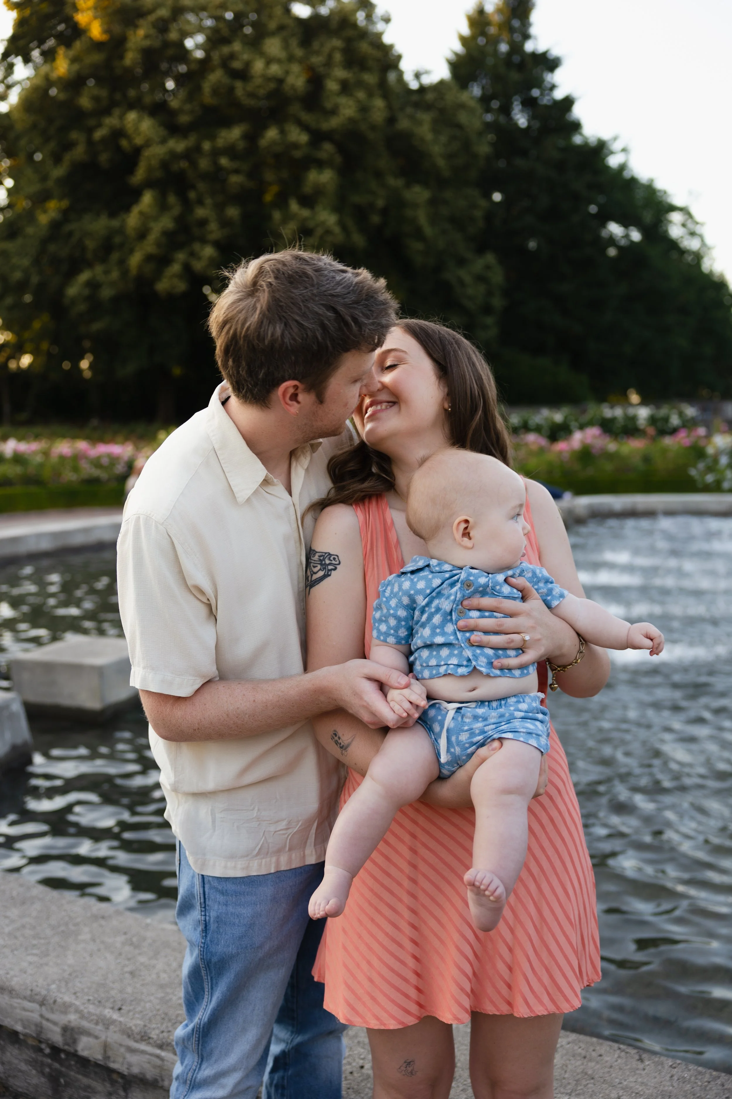 A young couple smiling and kissing while holding a baby near a body of water, with trees and flowers in the background.
