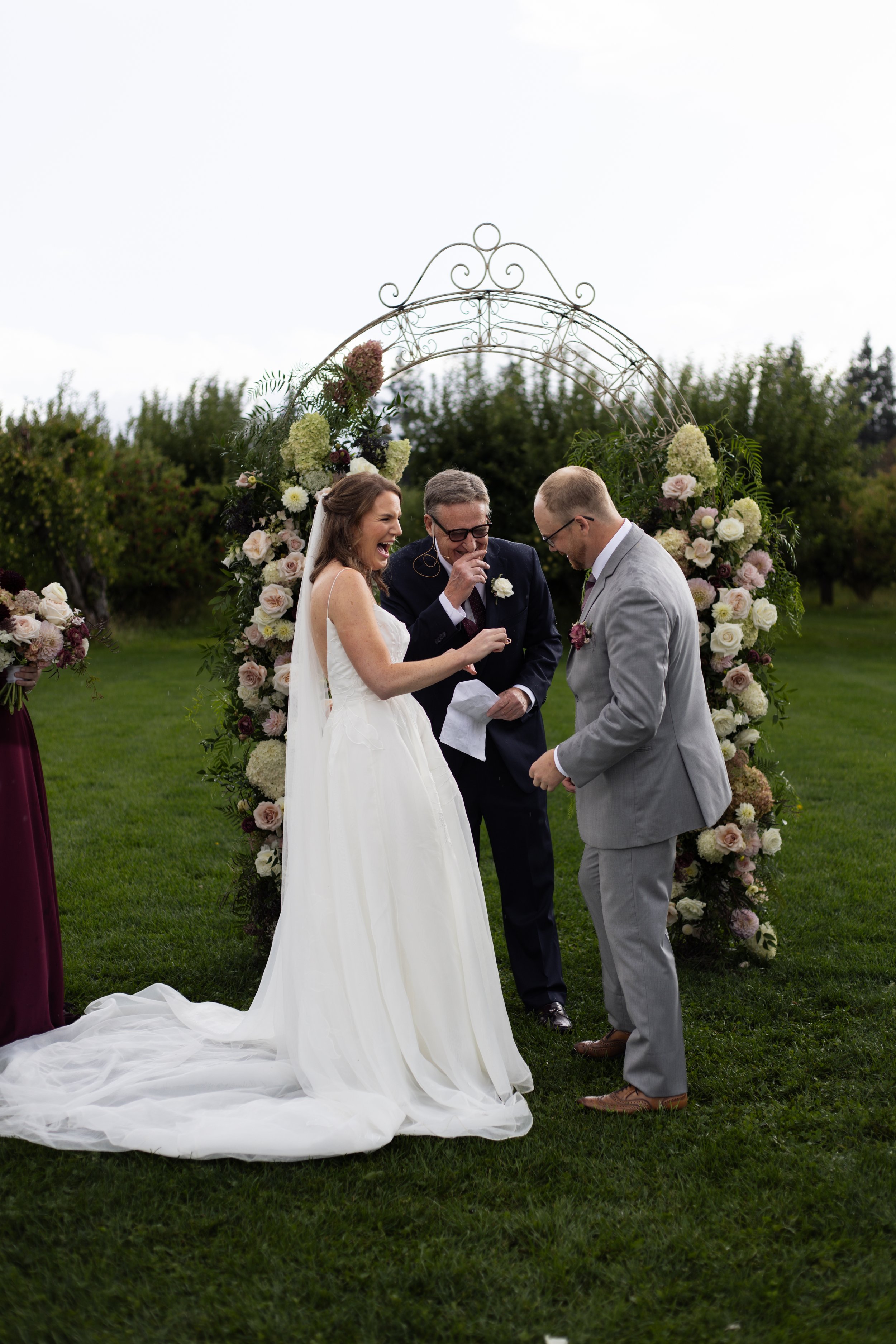 A wedding ceremony outdoors with a bride in a white gown and a groom in a light gray suit, exchanging vows under a floral arch with an officiant in a dark suit.