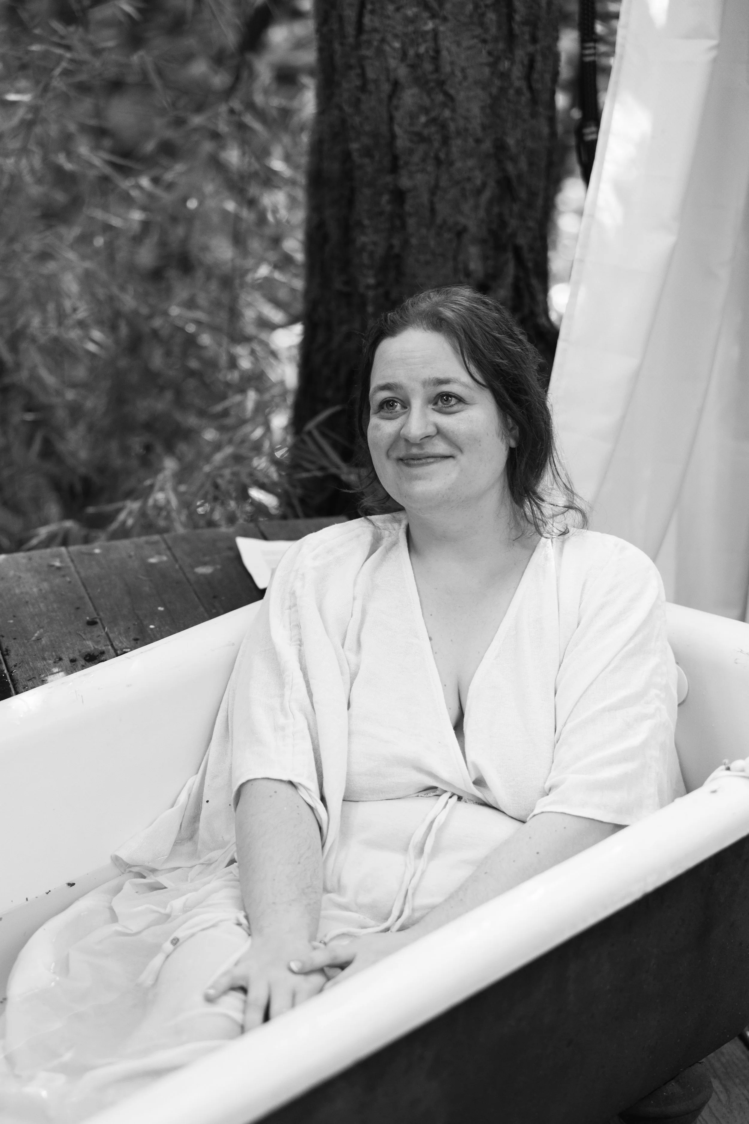 A woman with light skin and dark hair, dressed in a hospital gown, sitting in a bathtub, smiling.