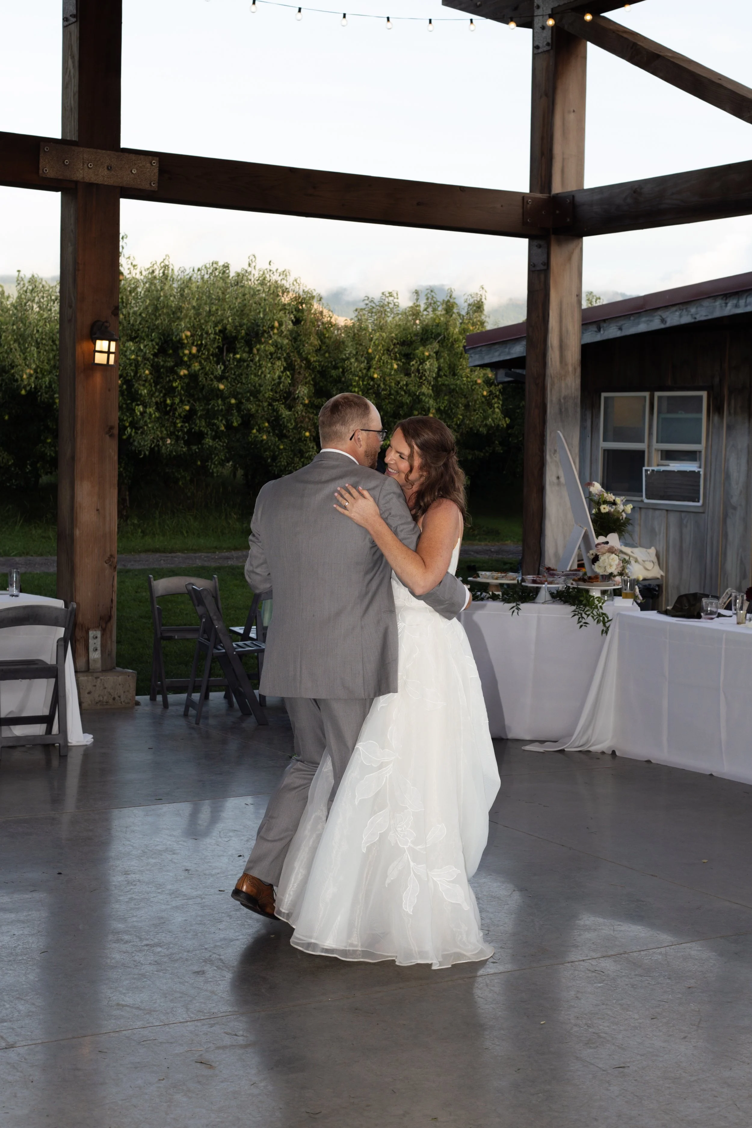A bride and groom sharing their first dance at an outdoor wedding reception, with trees and string lights in the background.