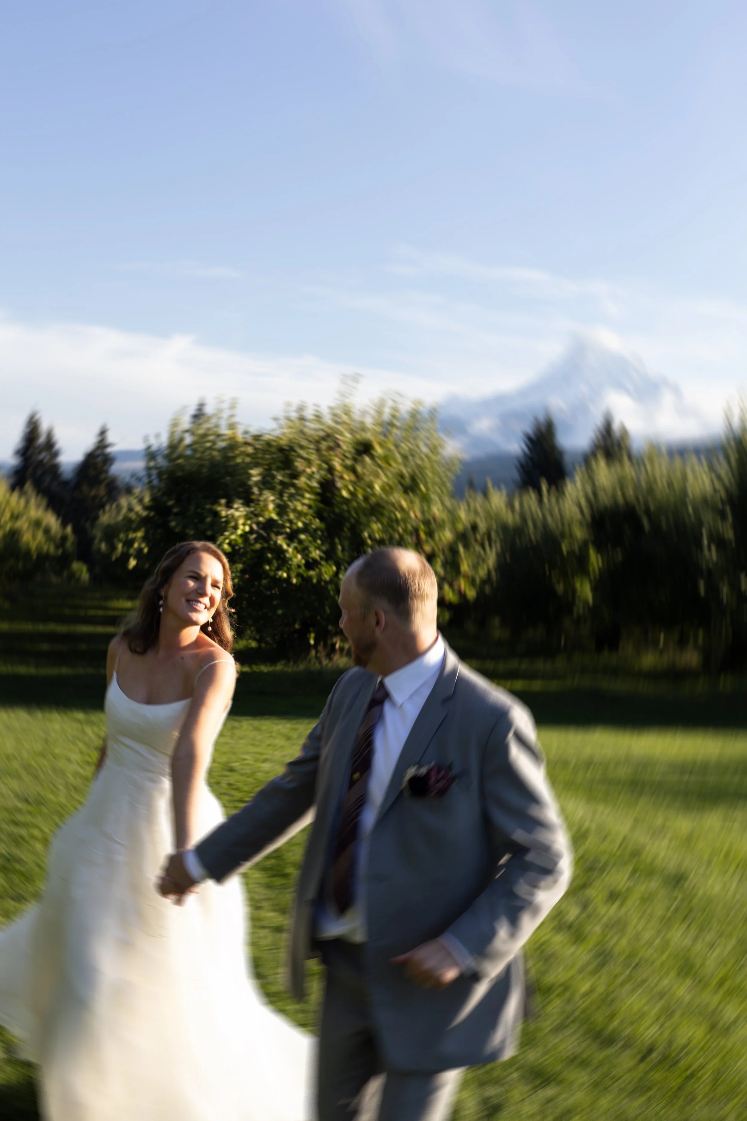 A bride and groom holding hands outdoors on a grassy area, with trees and a mountain in the background, during daylight.