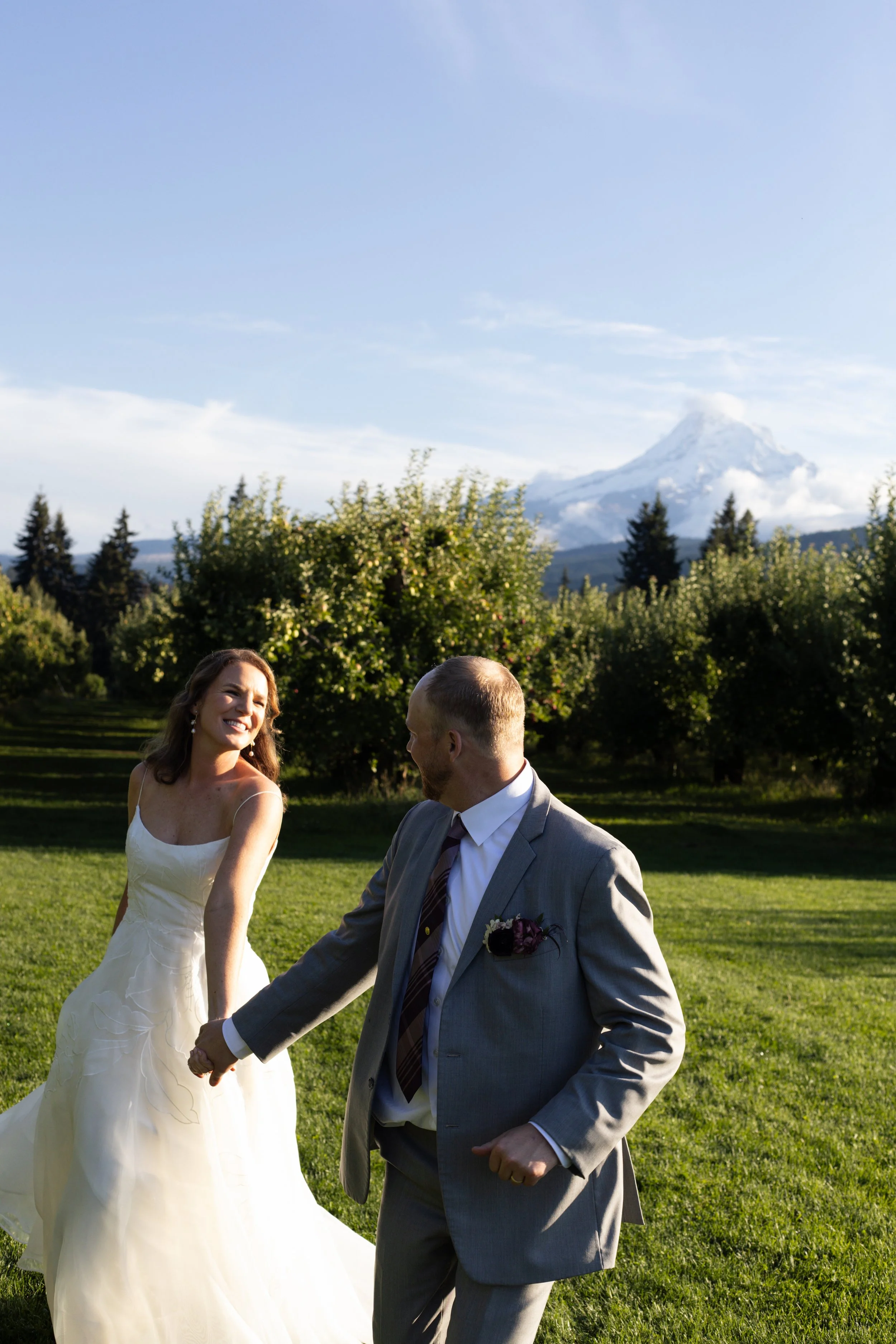 A bride and groom holding hands and dancing outdoors on a green lawn, with a mountainous landscape and snow-capped peak in the background.