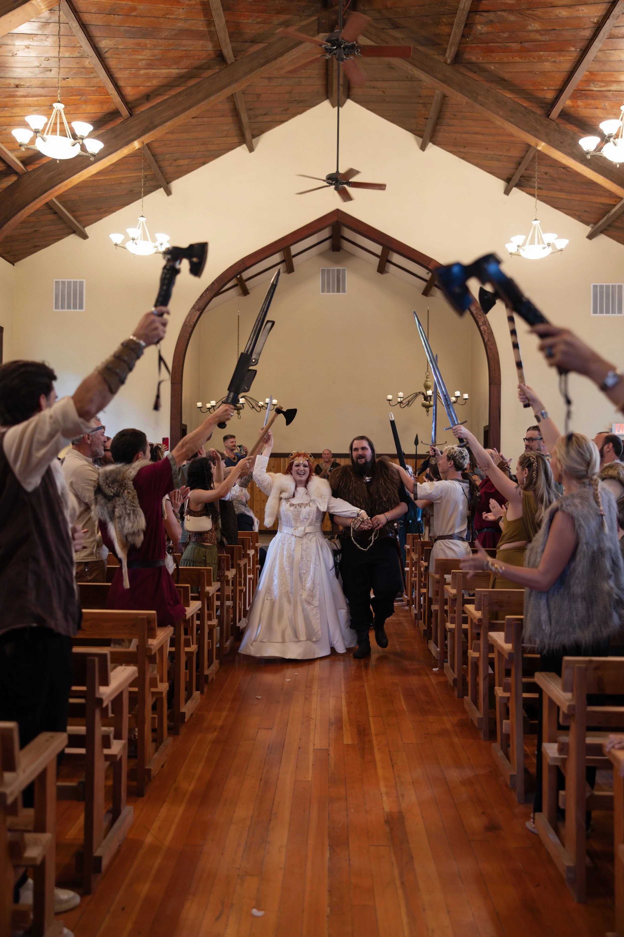 A wedding ceremony inside a wood-paneled hall with a high, vaulted ceiling. The bride and groom walk down the aisle, holding hands, surrounded by guests who are cheering and holding torches or light-emitting objects. The bride wears a white wedding d