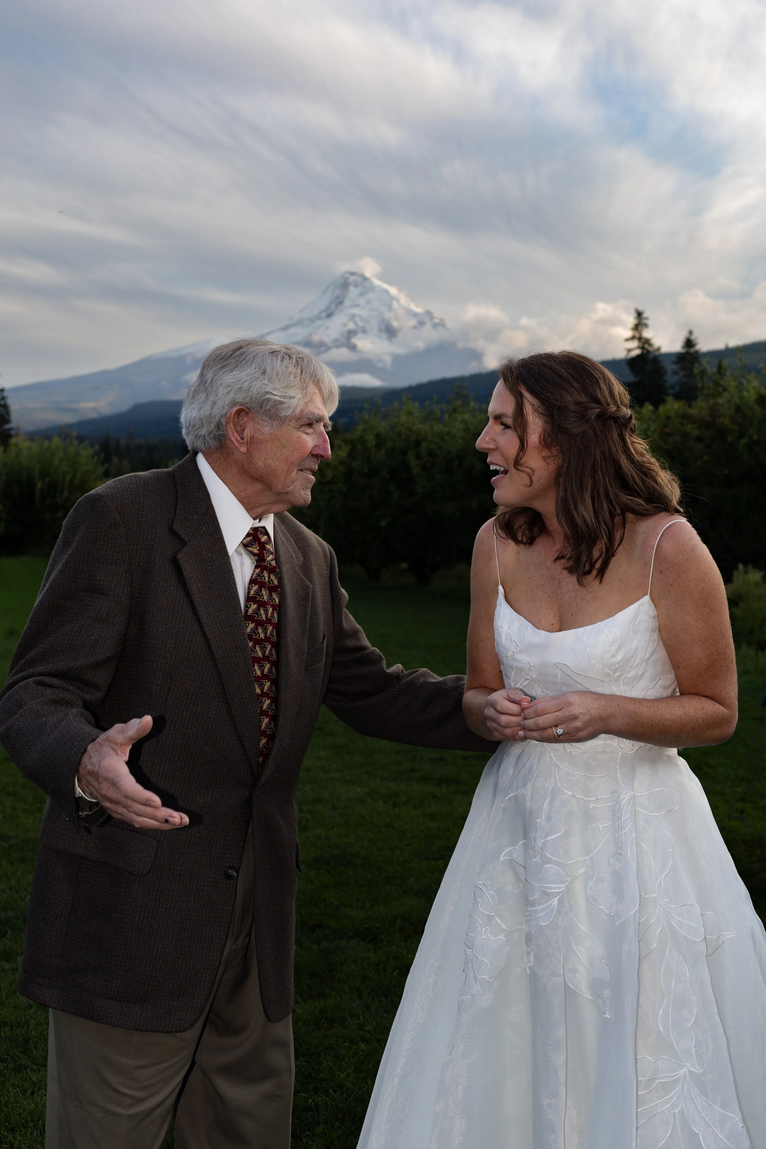 A senior man in a brown suit and a woman in a wedding dress at an outdoor wedding with a mountain and cloudy sky in the background.