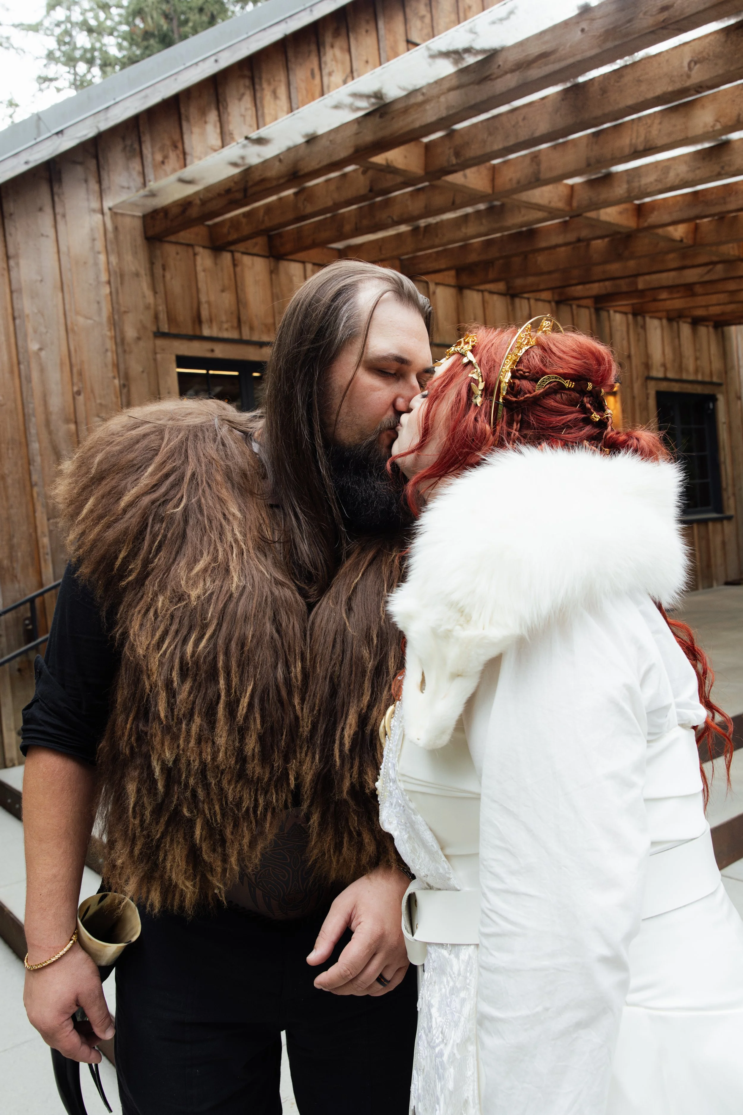 A man with long hair and a beard wearing a large fur coat is kissing a woman with red hair and a golden headpiece, dressed in a white outfit with a fur collar, in front of a wooden building.