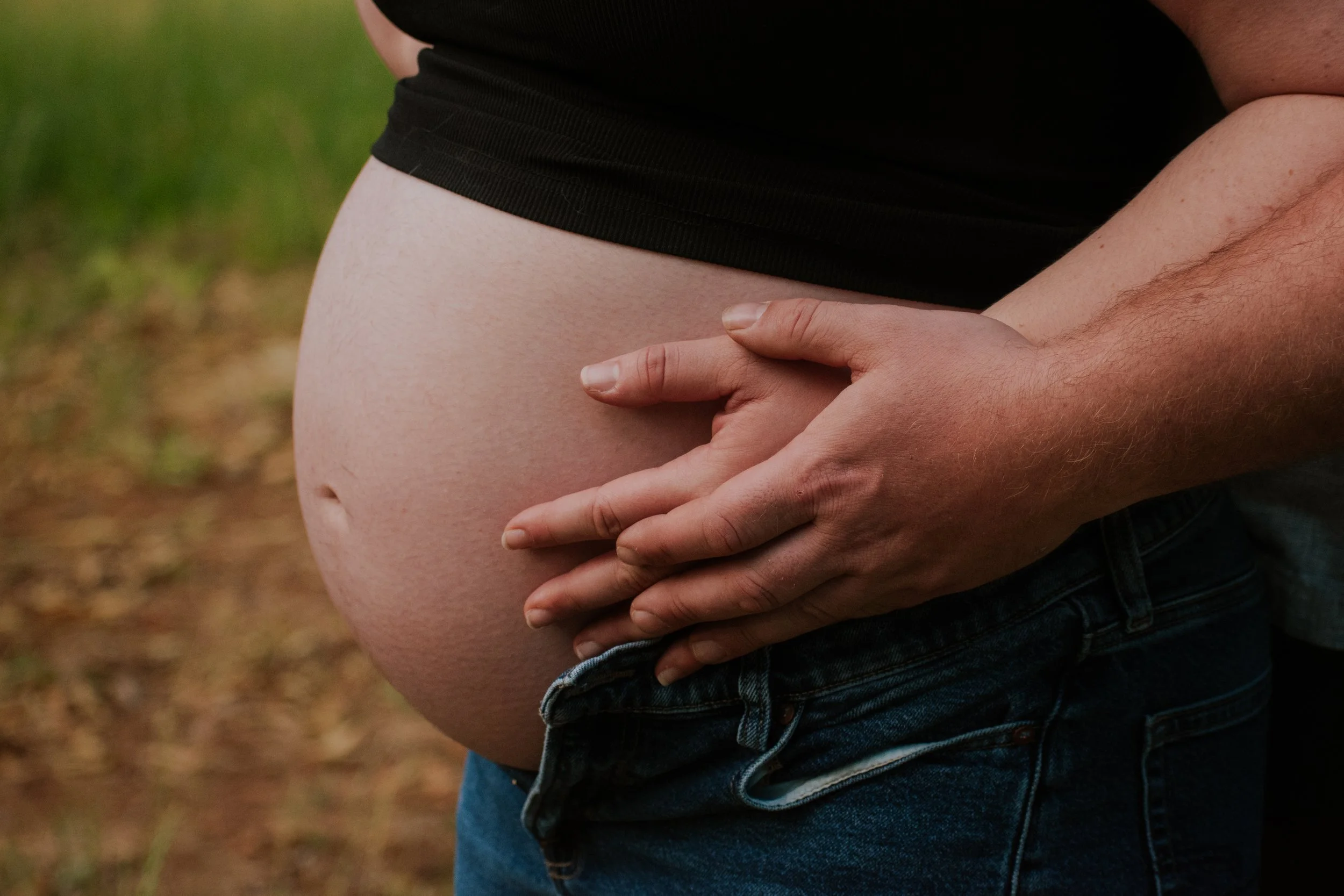 Close-up of an expecting woman with a man holding her baby bump from behind, outdoors in a natural setting.
