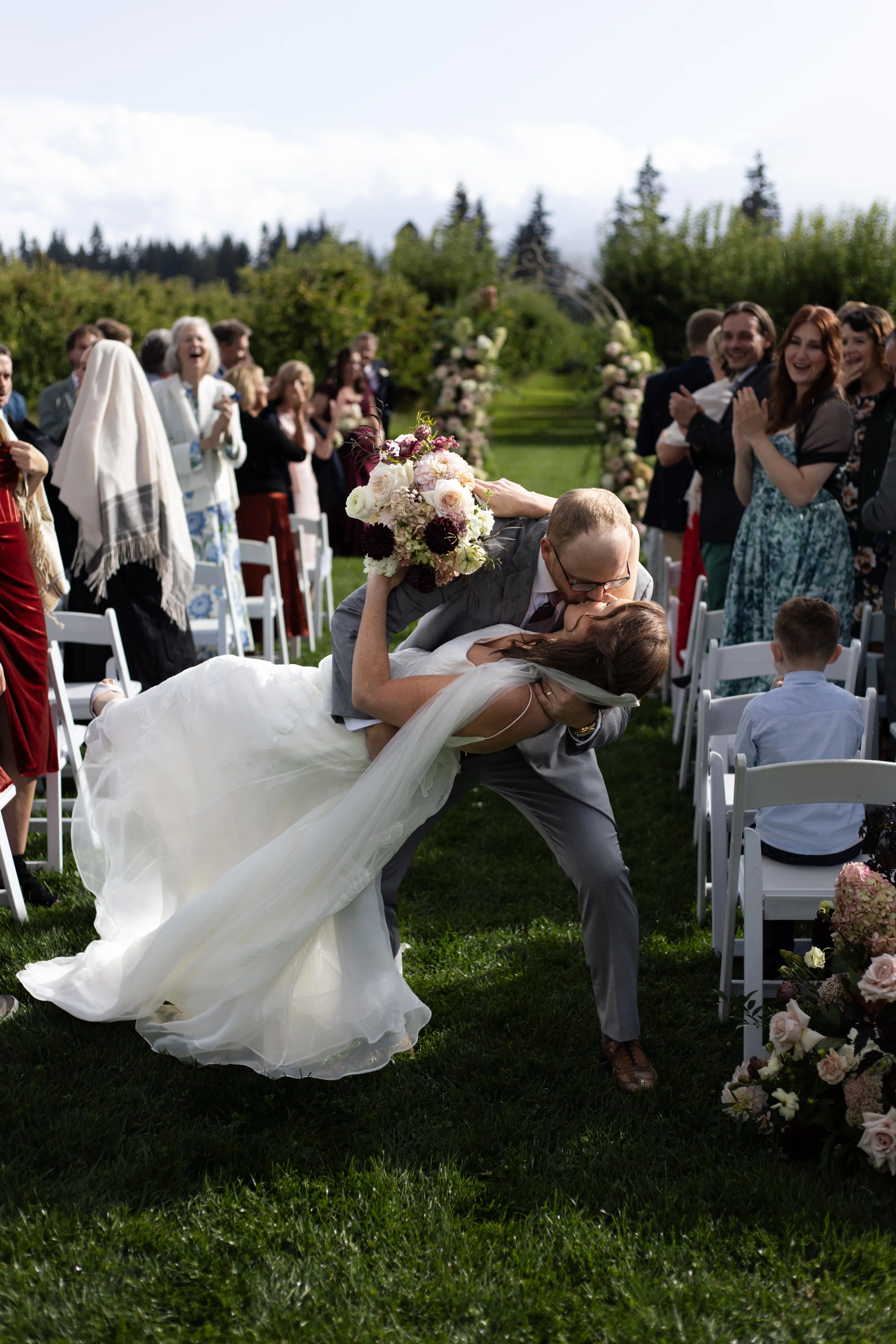 A bride and groom kissing during their outdoor wedding ceremony, with the groom helping the bride bend backward amid guests and floral arrangements.