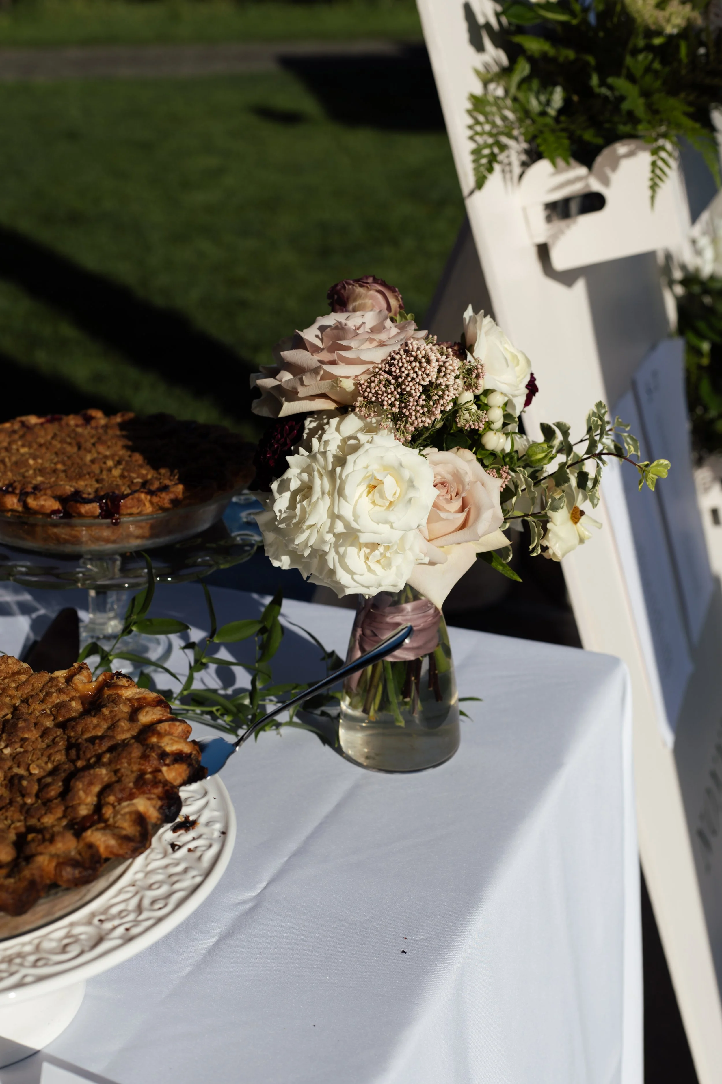 A floral centerpiece with white and blush roses, small pink flowers, and greenery in a glass vase on a white table, with a tray of baked goods nearby and an outdoor background.