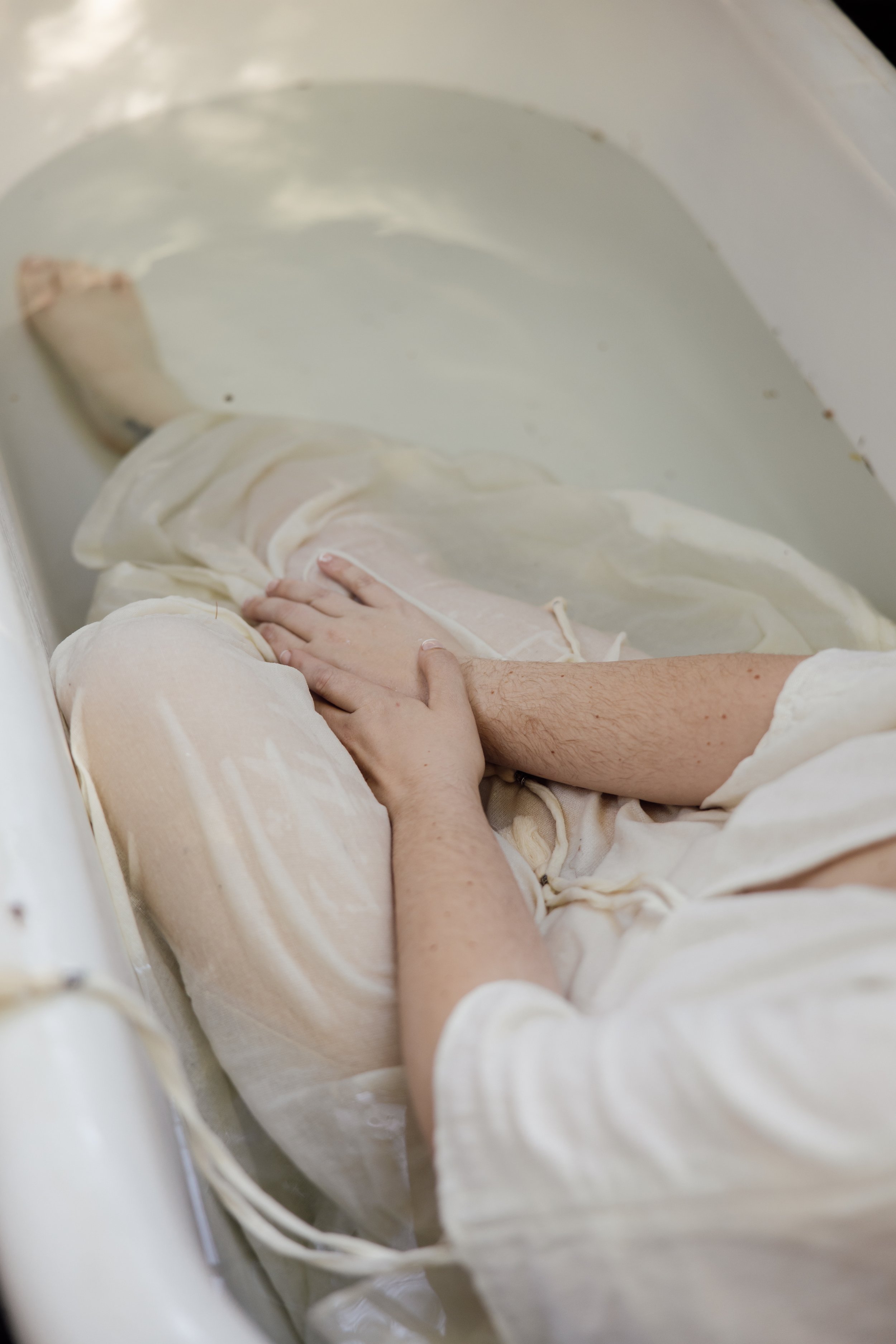 A baby lying in a white bassinet with its hand resting on a white blanket, and an adult's hand gently touching the baby's arm.