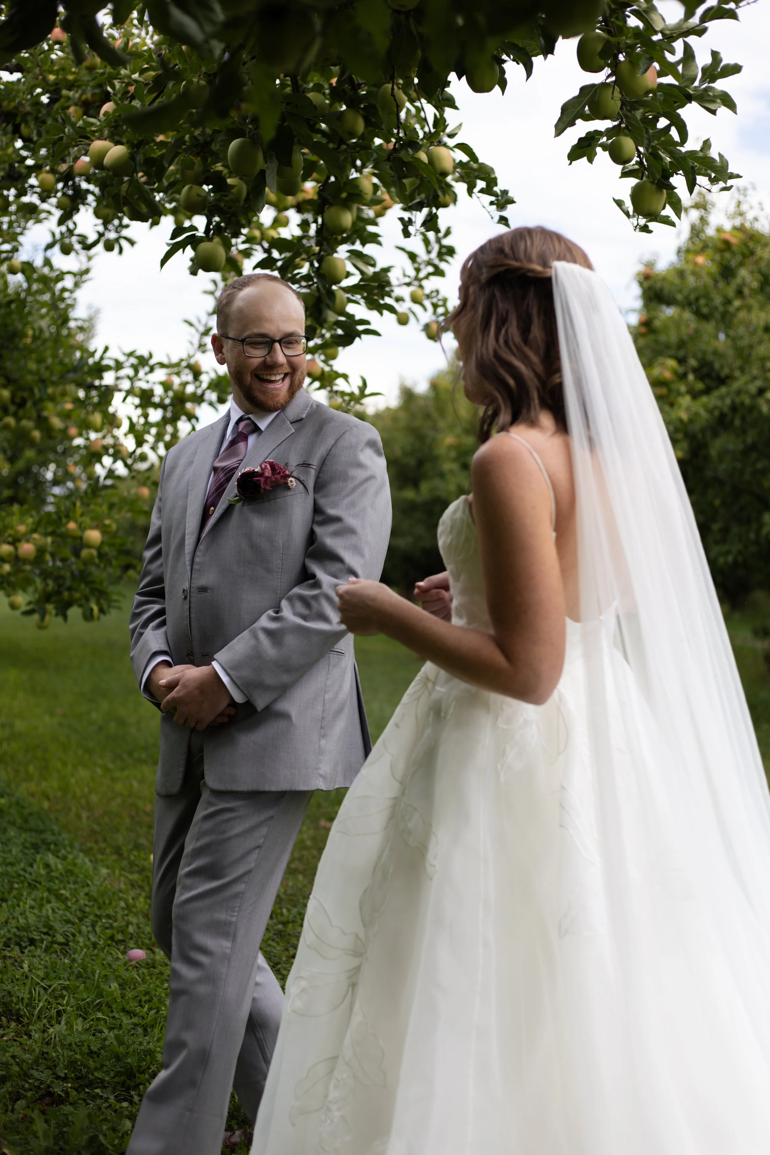 A couple is outdoors during their wedding ceremony, with a man in a gray suit and a woman in a white wedding dress and veil standing under an apple tree, smiling and holding hands.