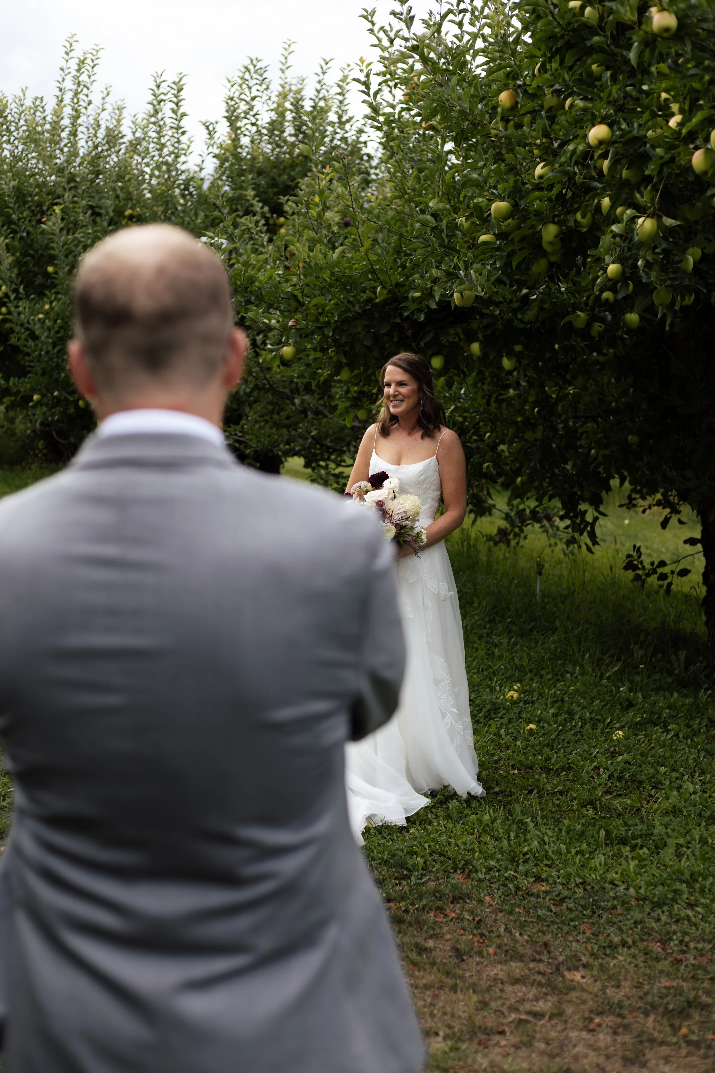 A bride stands outdoors holding a bouquet, while a man in a gray suit stands with his back to the camera, in front of an apple tree with green apples.