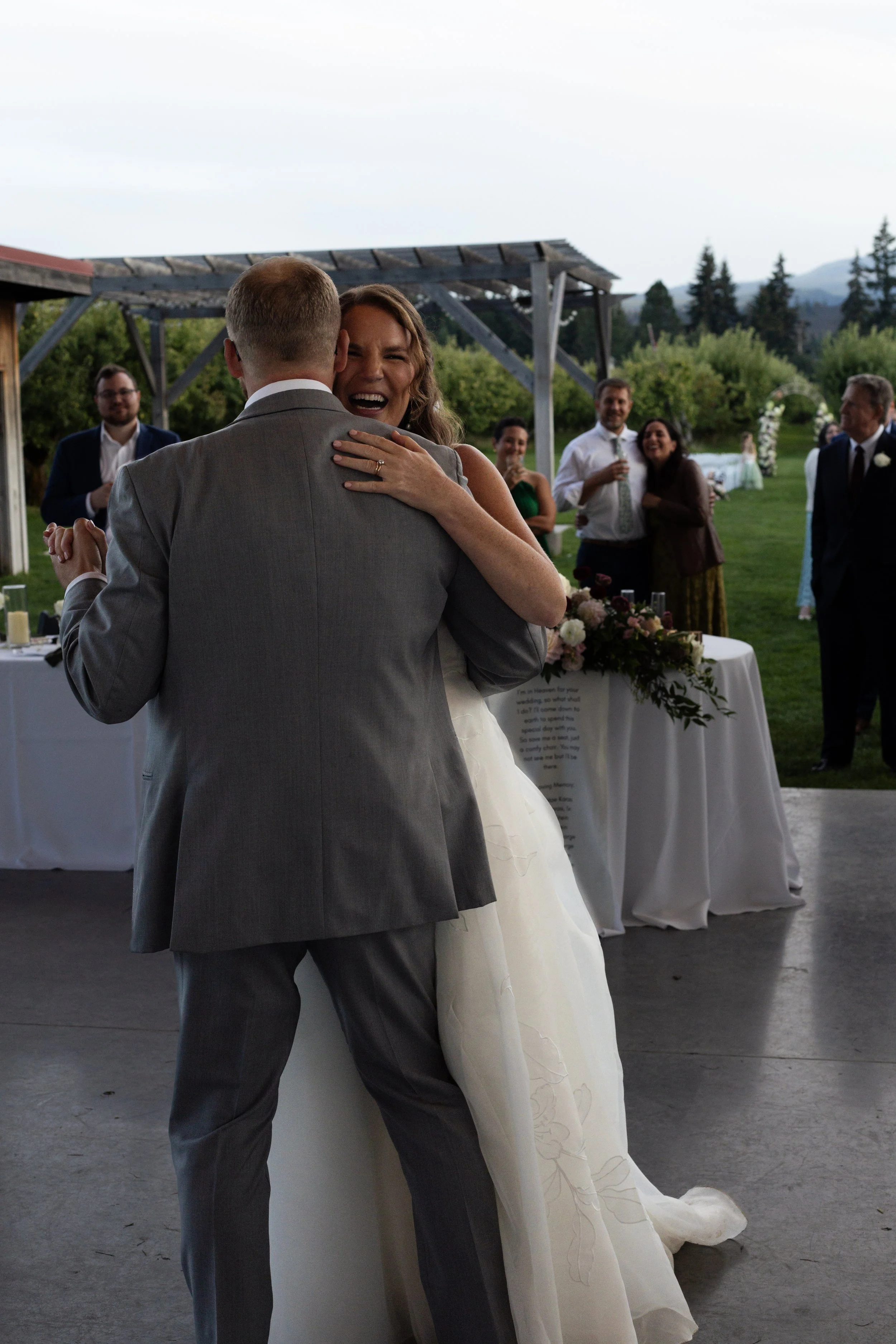 A bride and groom share a dance at an outdoor wedding reception, with guests watching and smiling in the background.