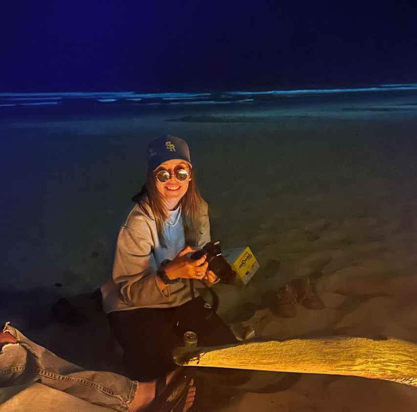 A woman sitting on the beach at night, holding a camera and smiling at the camera, with a headlamp and a pair of shoes nearby.