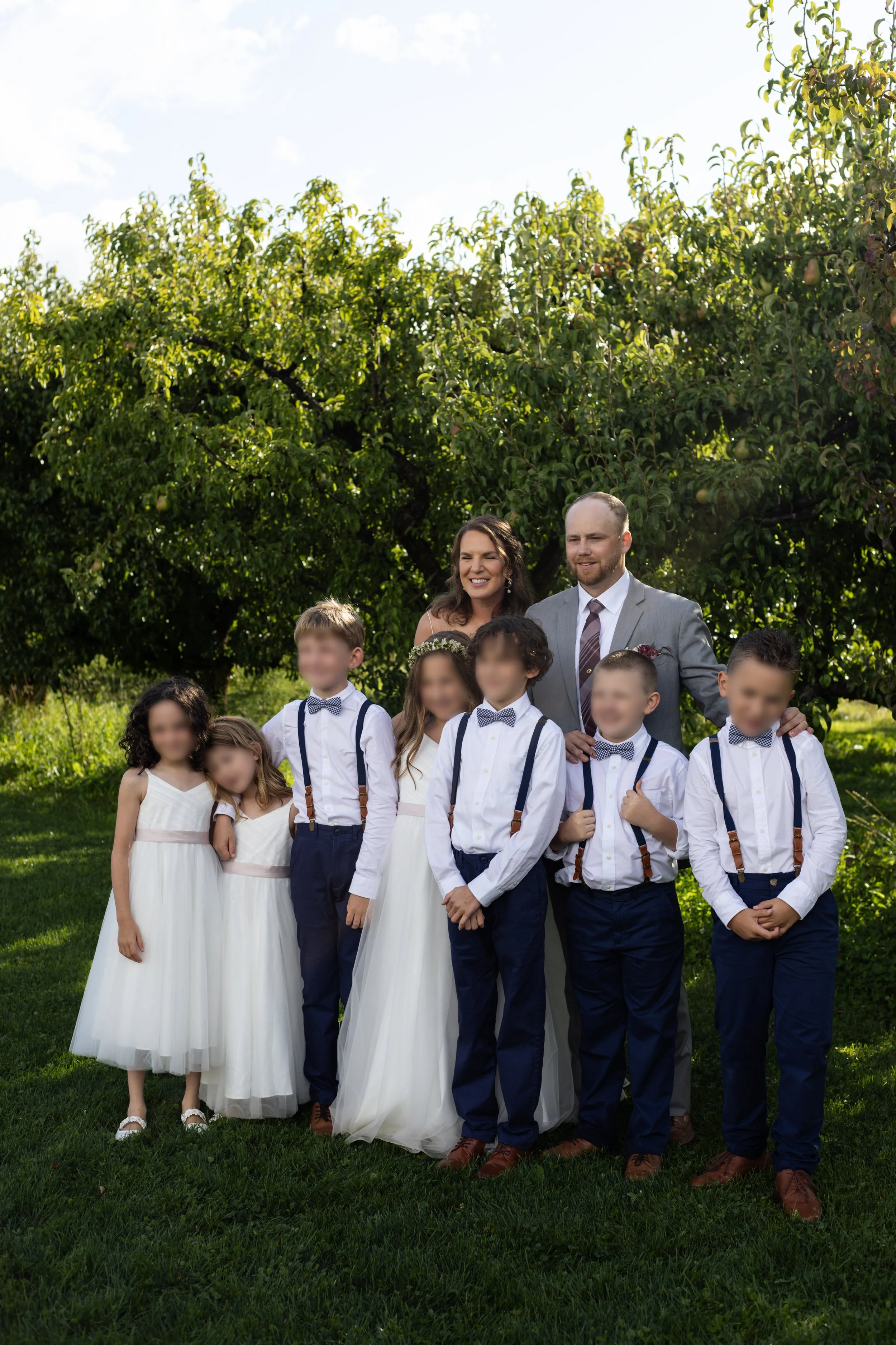 A group of children dressed in formal wedding attire with a woman and man, outdoors in front of green trees, on a sunny day.