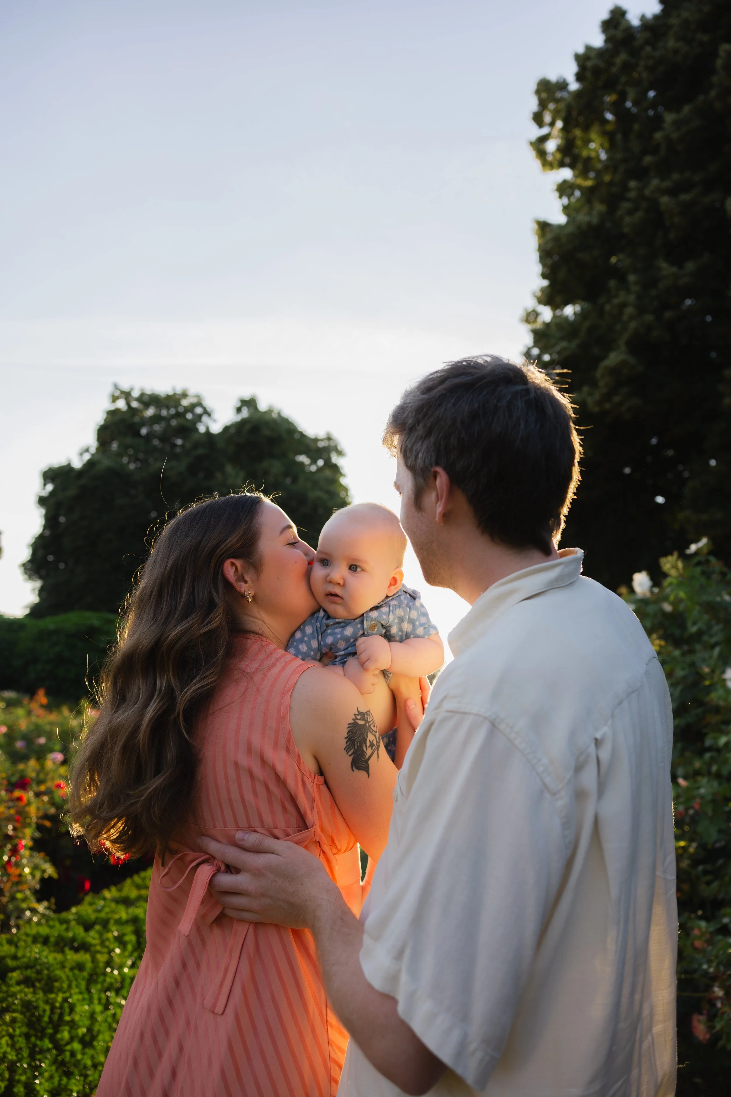 A woman holding a baby and a man is facing each other in a park during sunset, with trees and flowers in the background.