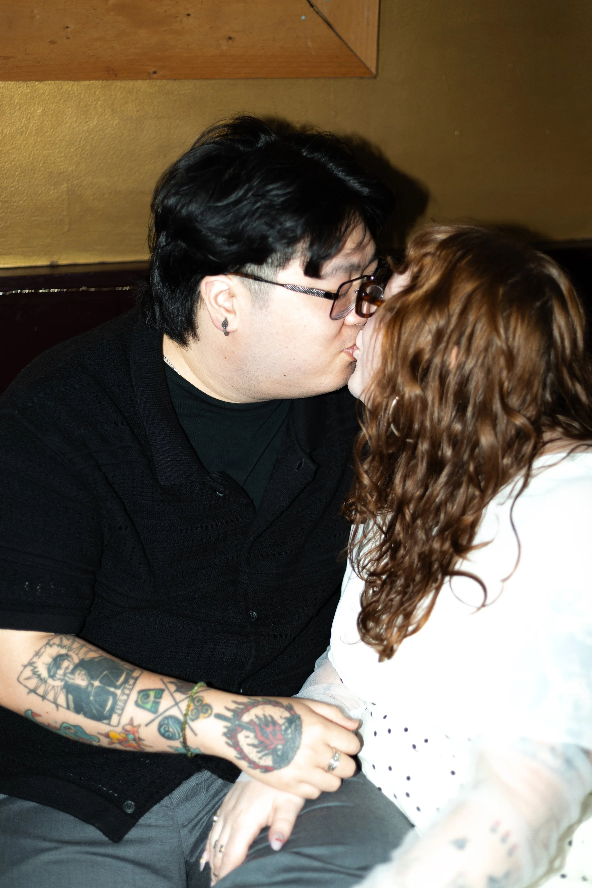 A couple sharing a kiss, with the woman having curly red hair and tattoos on her arm, and the man wearing glasses and earrings, in an indoor setting.
