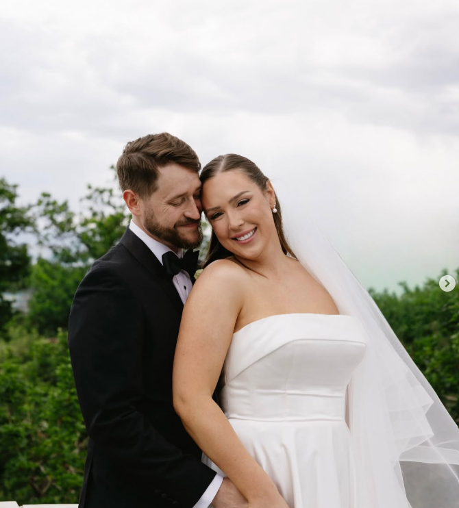 A newlywed couple on their wedding day outdoors, the groom in a black tuxedo with a bow tie and the bride in a strapless white wedding gown, smiling affectionately.