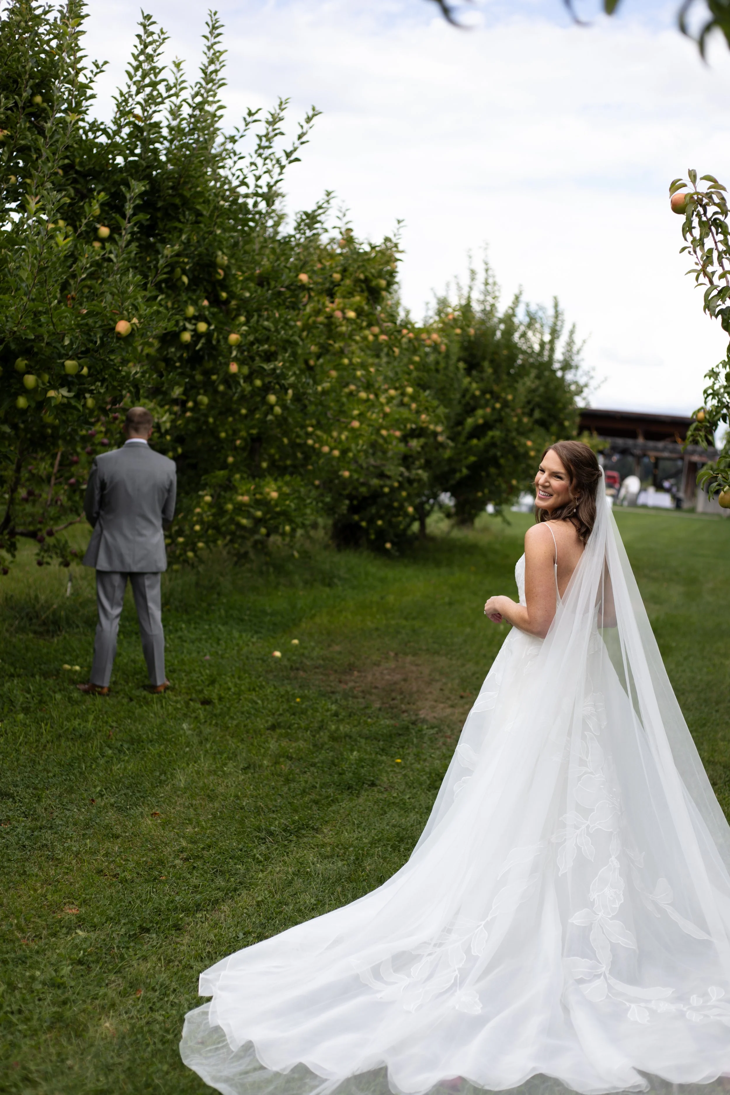 A bride in a white wedding gown with a long train, smiling and looking over her shoulder, standing in a grassy orchard with apple trees, while a groom in a gray suit faces away in the background.