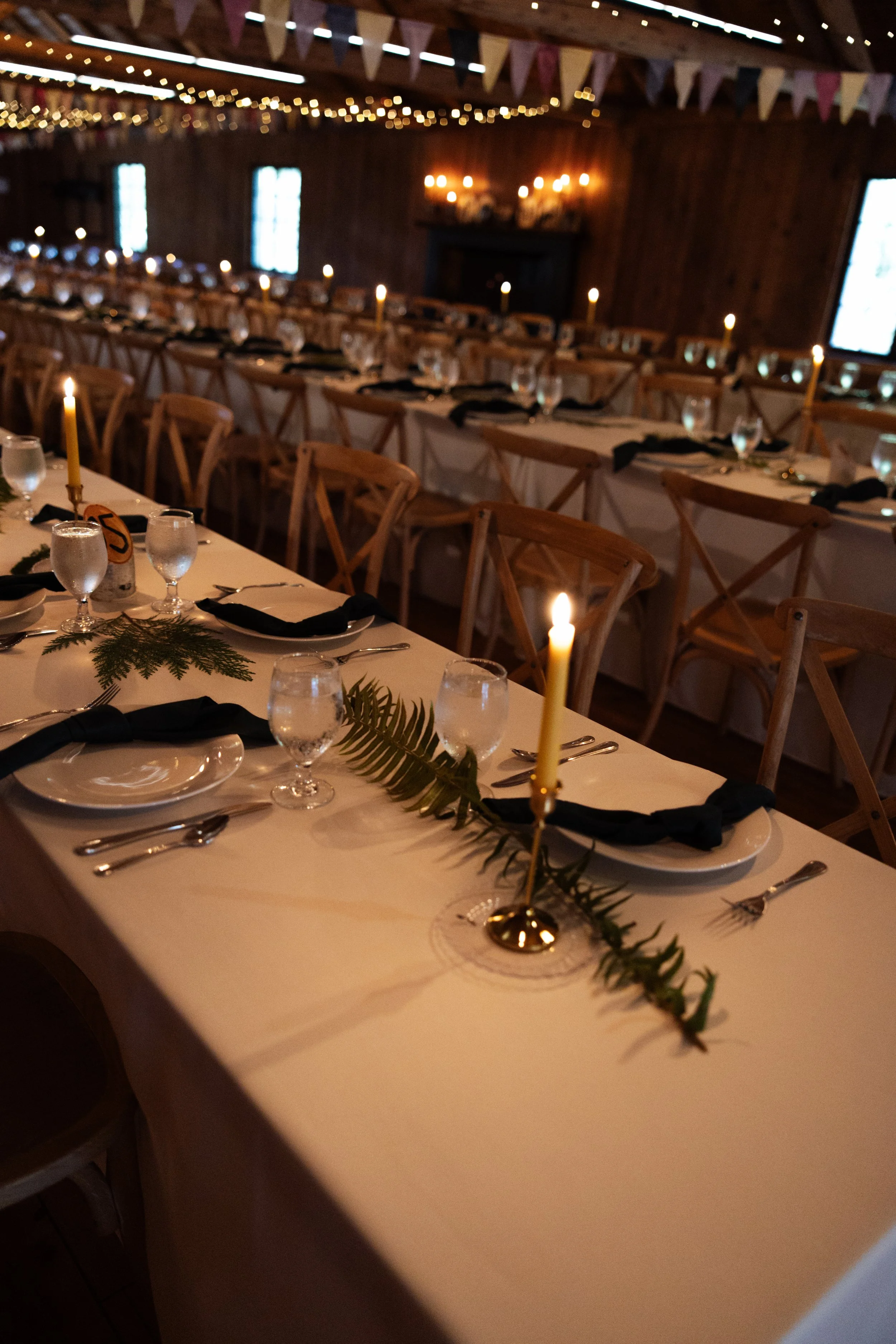 A long banquet table set for a celebration with white tablecloths, black napkins, wine glasses, and plates. The table is decorated with green foliage and candles. The room has wooden walls, string lights, bunting, and a fireplace in the background.
