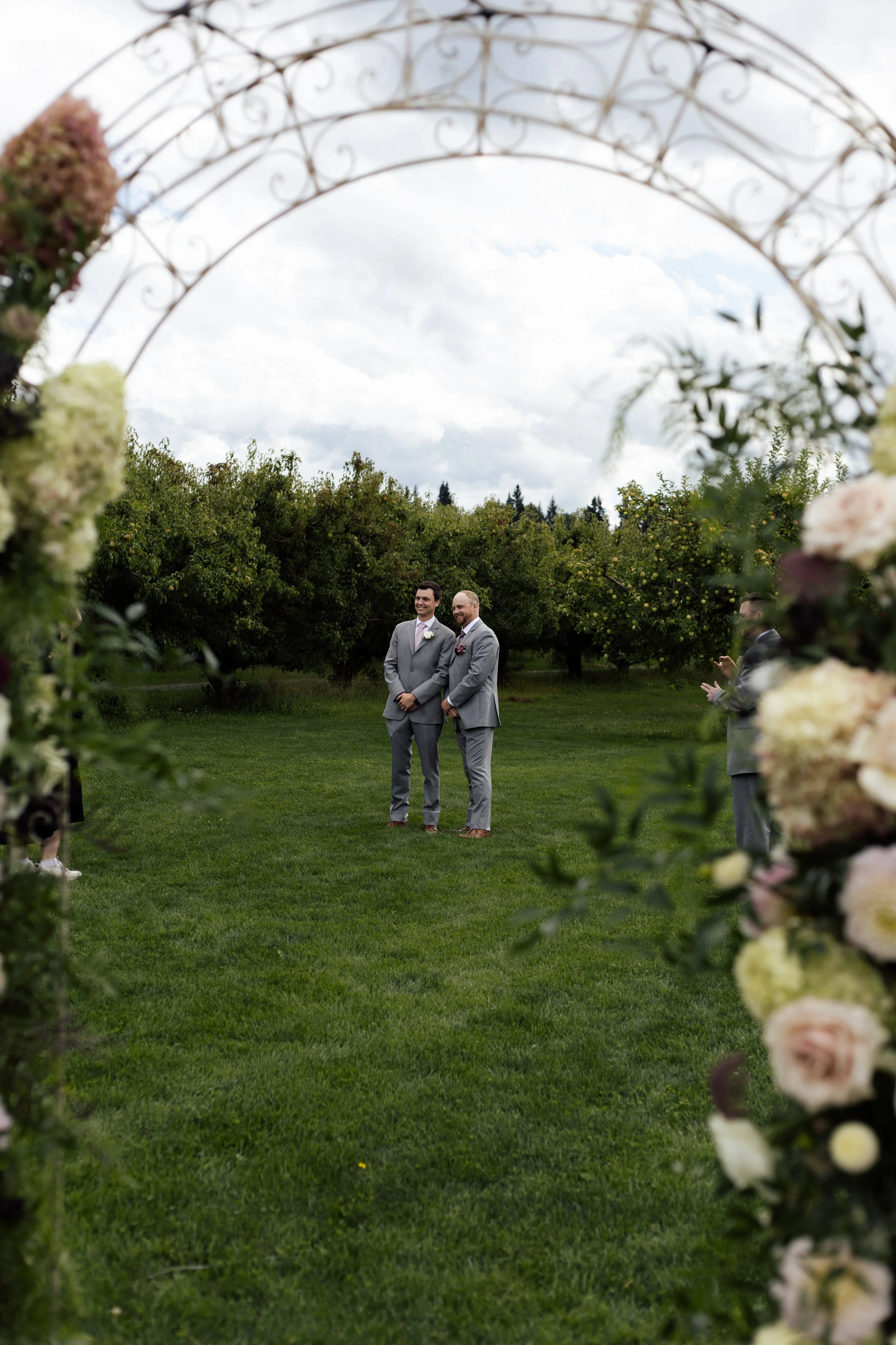 Two men in suits standing outdoors in a garden or park, framed by a decorative floral archway. It is daytime with cloudy skies, and there are trees and a grassy area in the background.