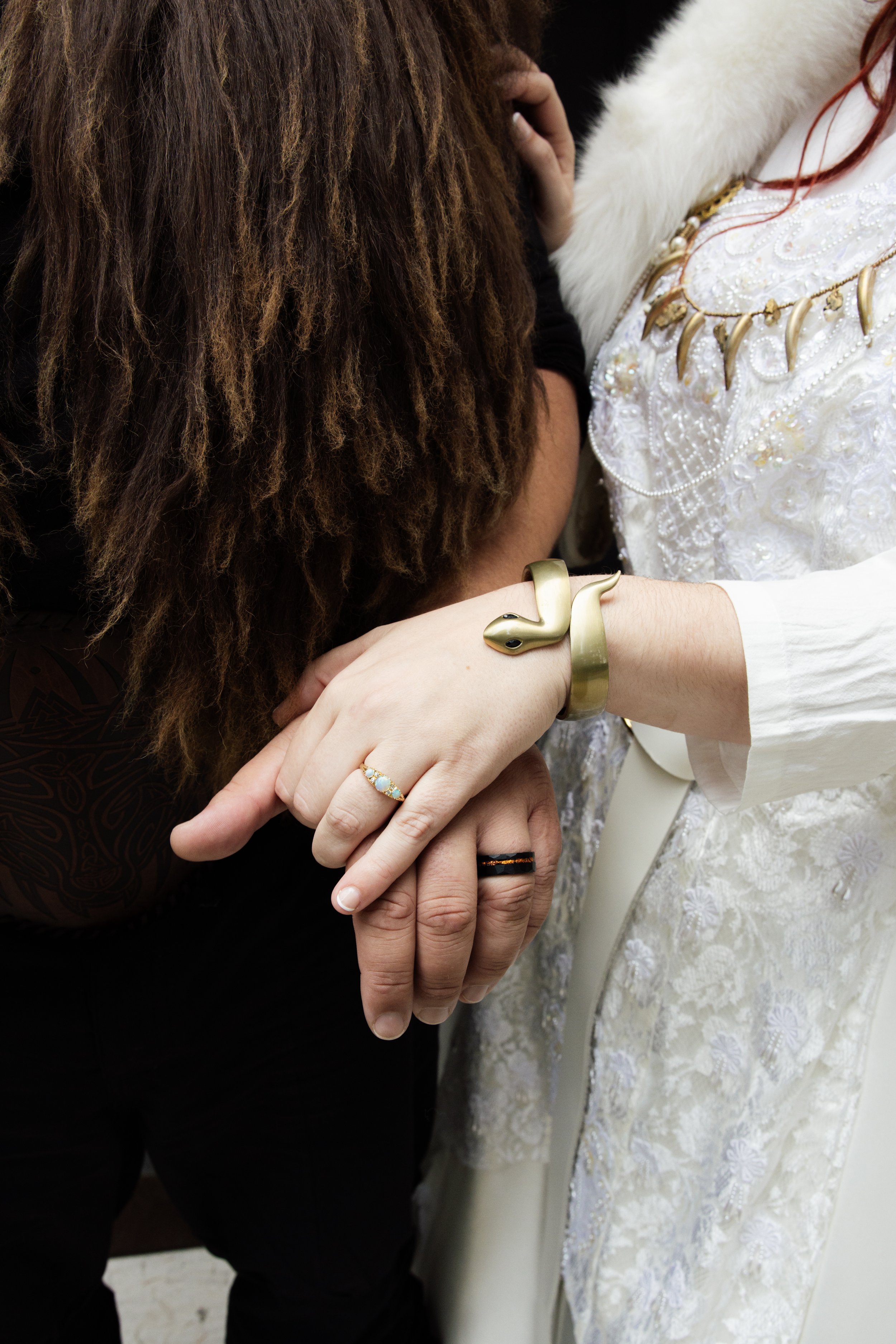 A close-up of a group of people holding hands, with one wearing a gold snake bracelet and another wearing a ring with opalescent stones. The background reveals part of a white dress with floral embroidery and a woman with long red hair. The setting a